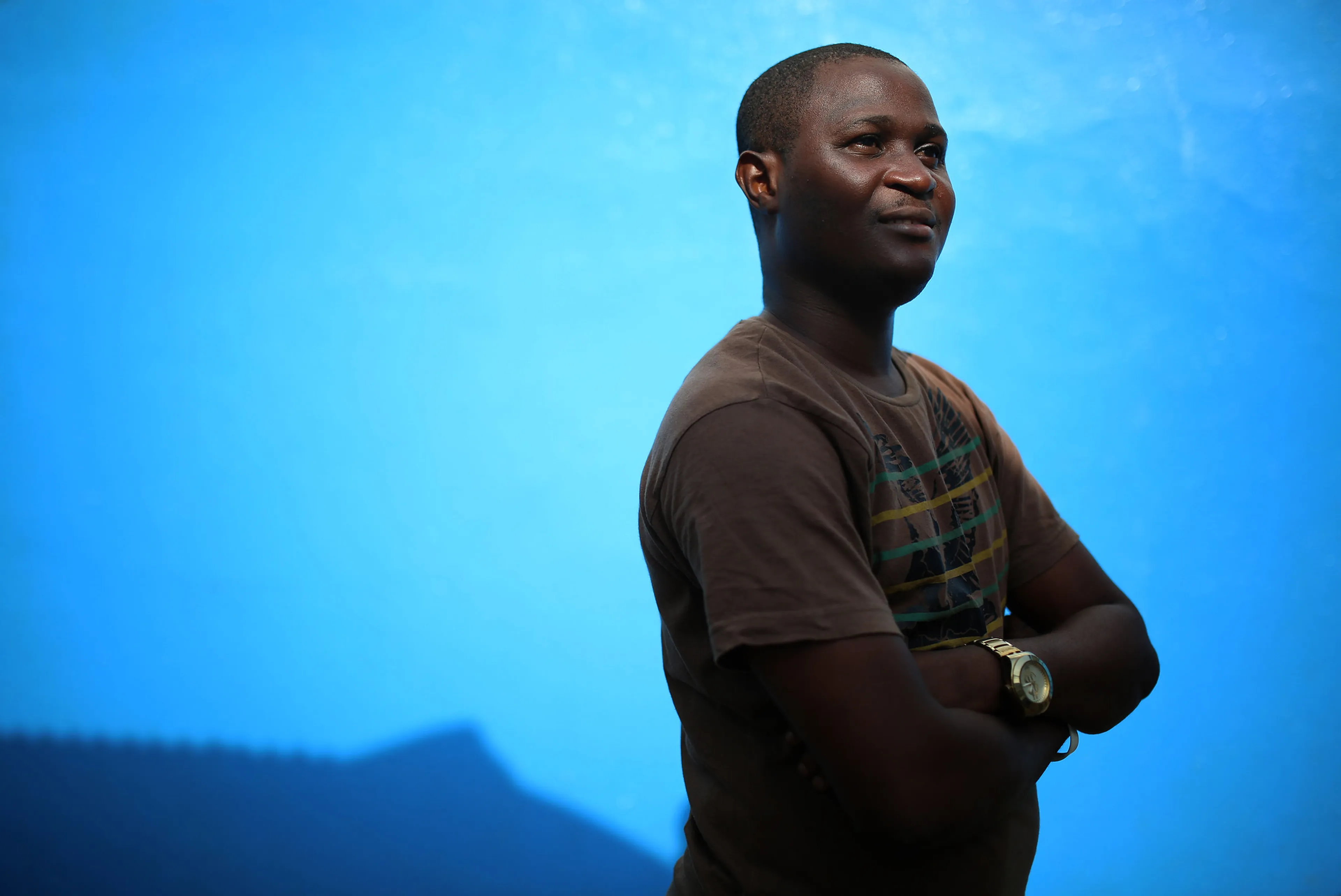 Person in a brown shirt with crossed arms, standing against a bright blue background, looking confidently to the side.