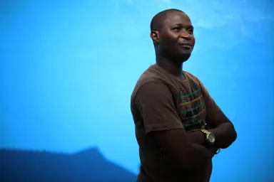 Person in a brown shirt with crossed arms, standing against a bright blue background, looking confidently to the side.