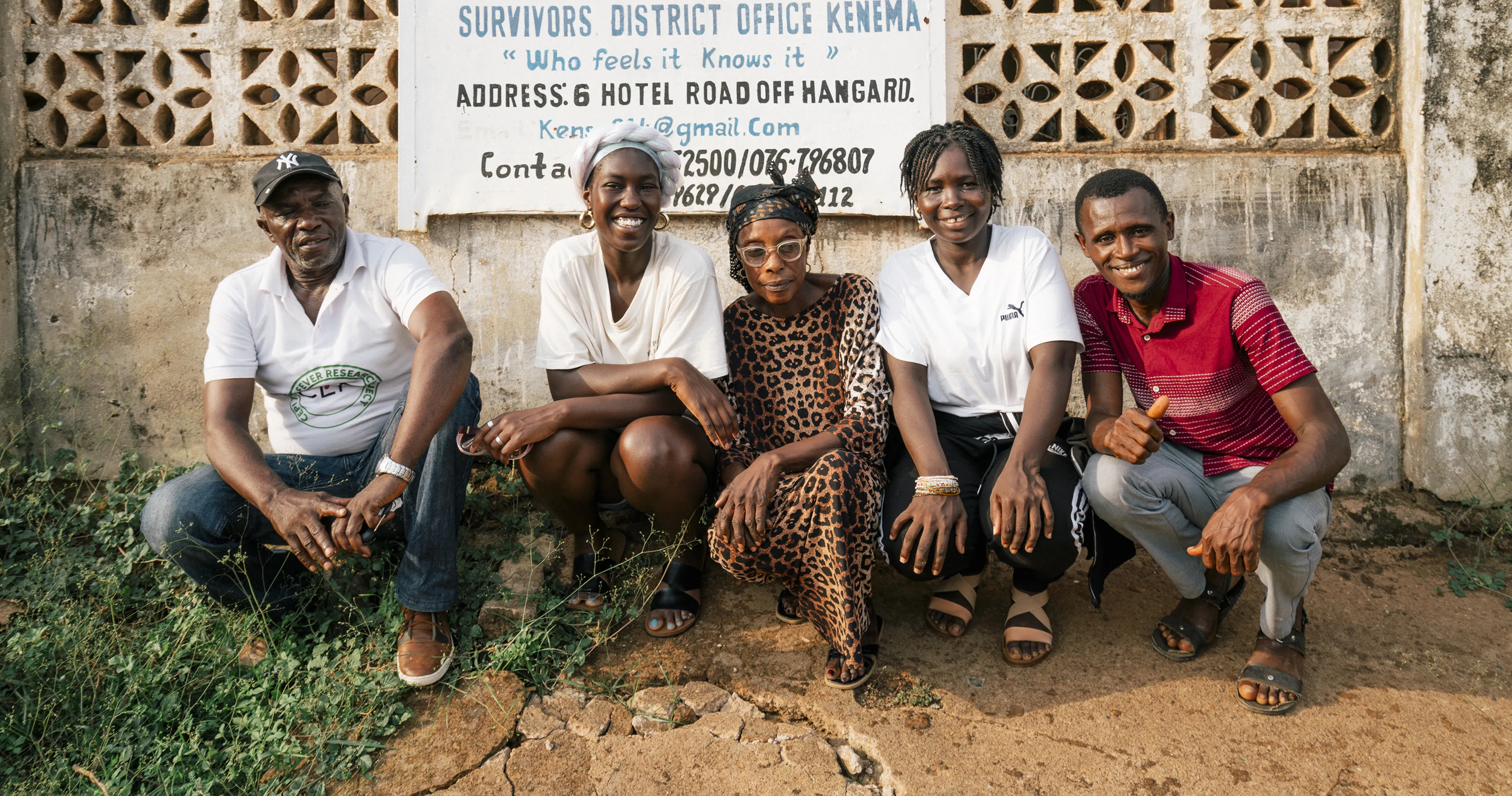 Five people sitting and smiling in front of a wall with a sign for the Sierra Leone Association of Ebola Survivors in Kenema.