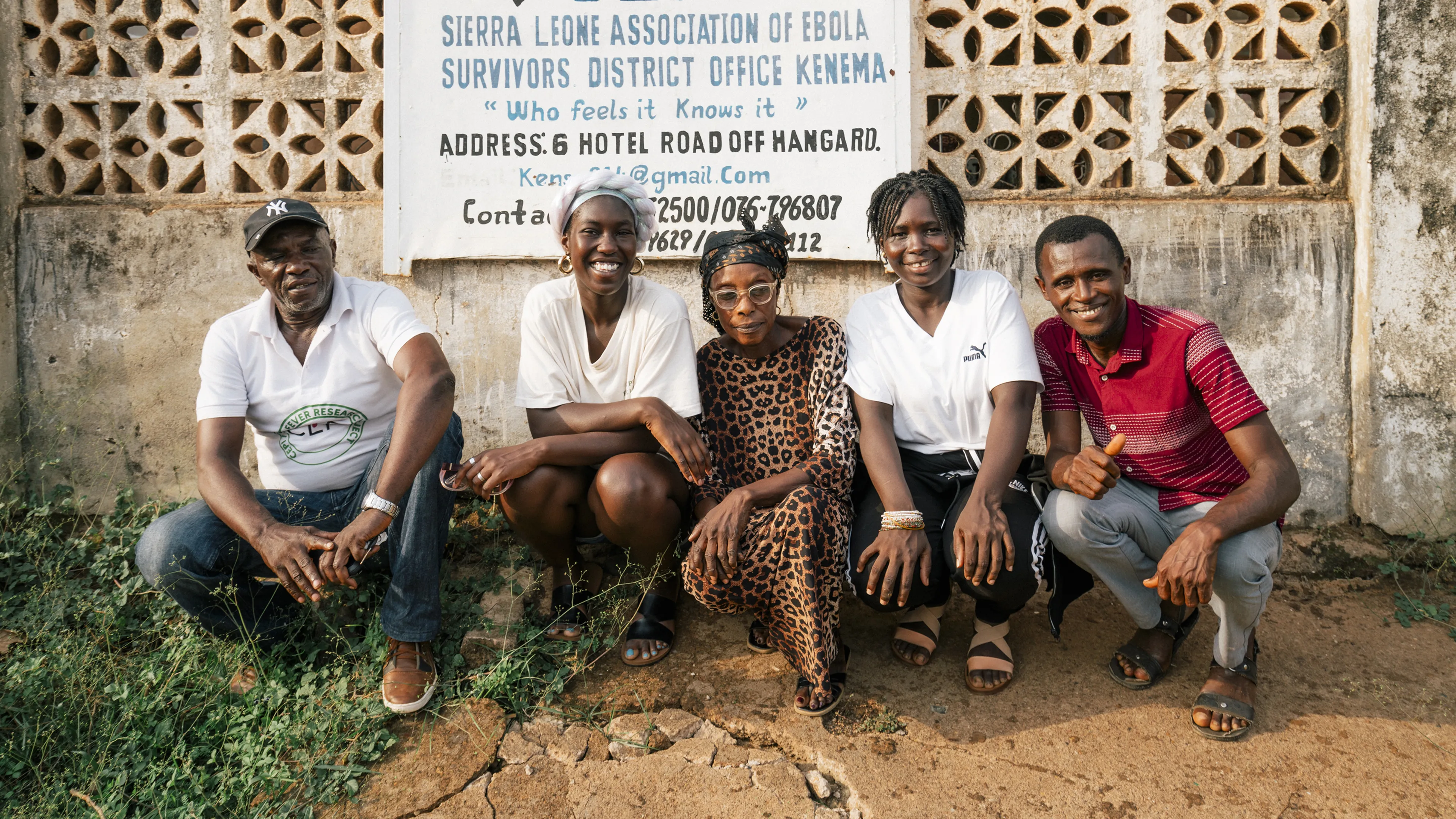 Five people sitting and smiling in front of a wall with a sign for the Sierra Leone Association of Ebola Survivors in Kenema.