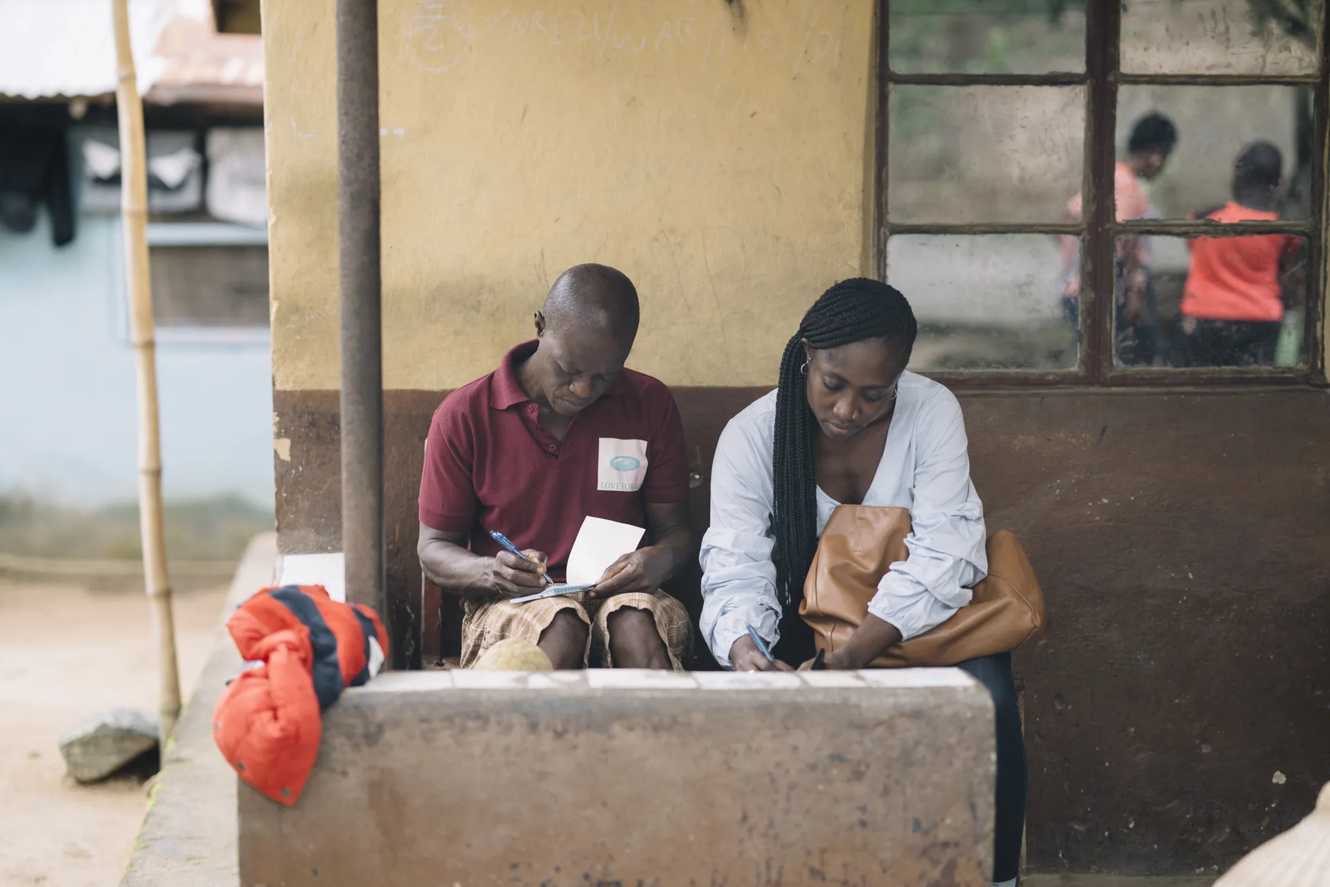 Two people sit on a concrete bench, writing on paper, with a yellow and brown wall behind them and a window to the side.