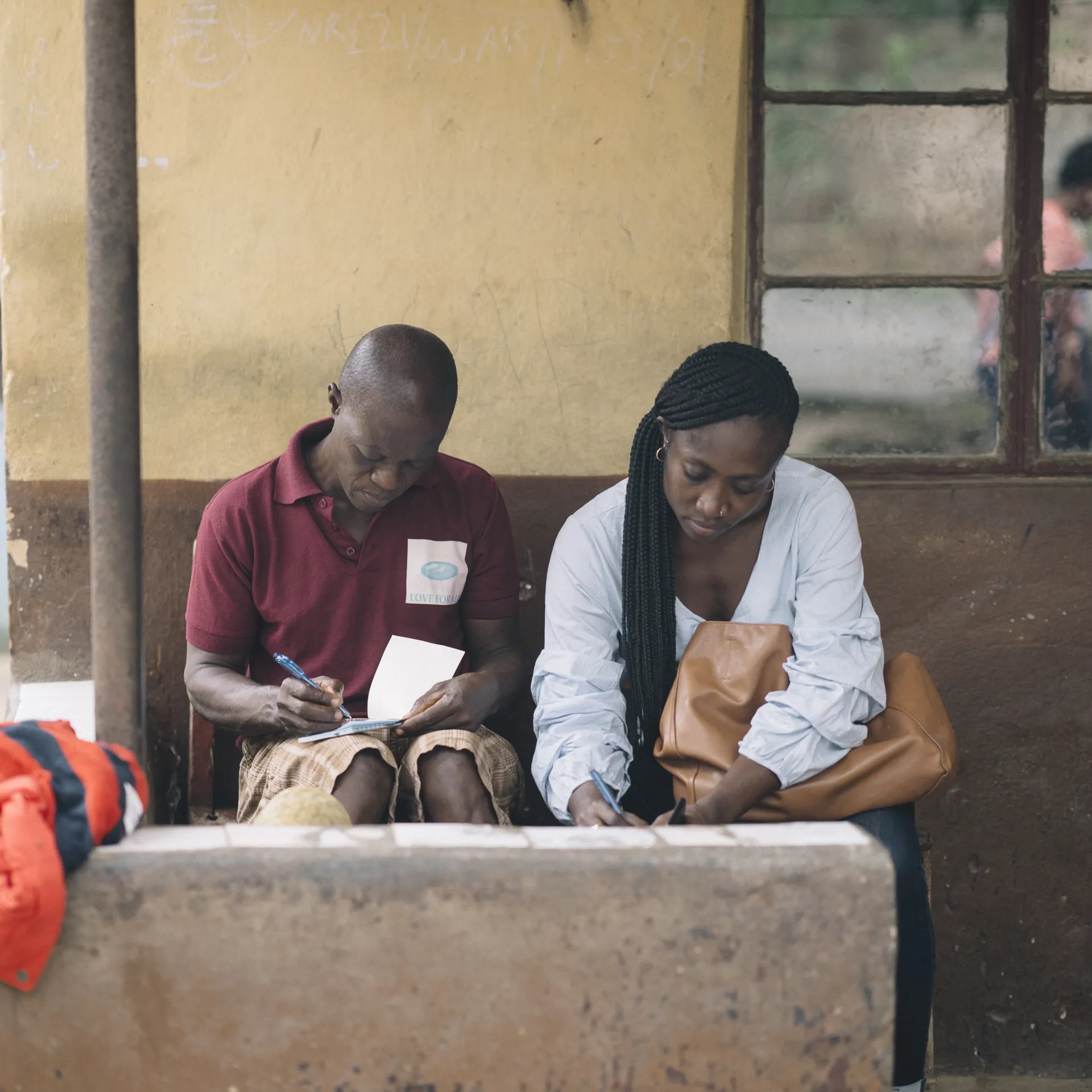 Two people sit on a concrete bench, writing on paper, with a yellow and brown wall behind them and a window to the side.