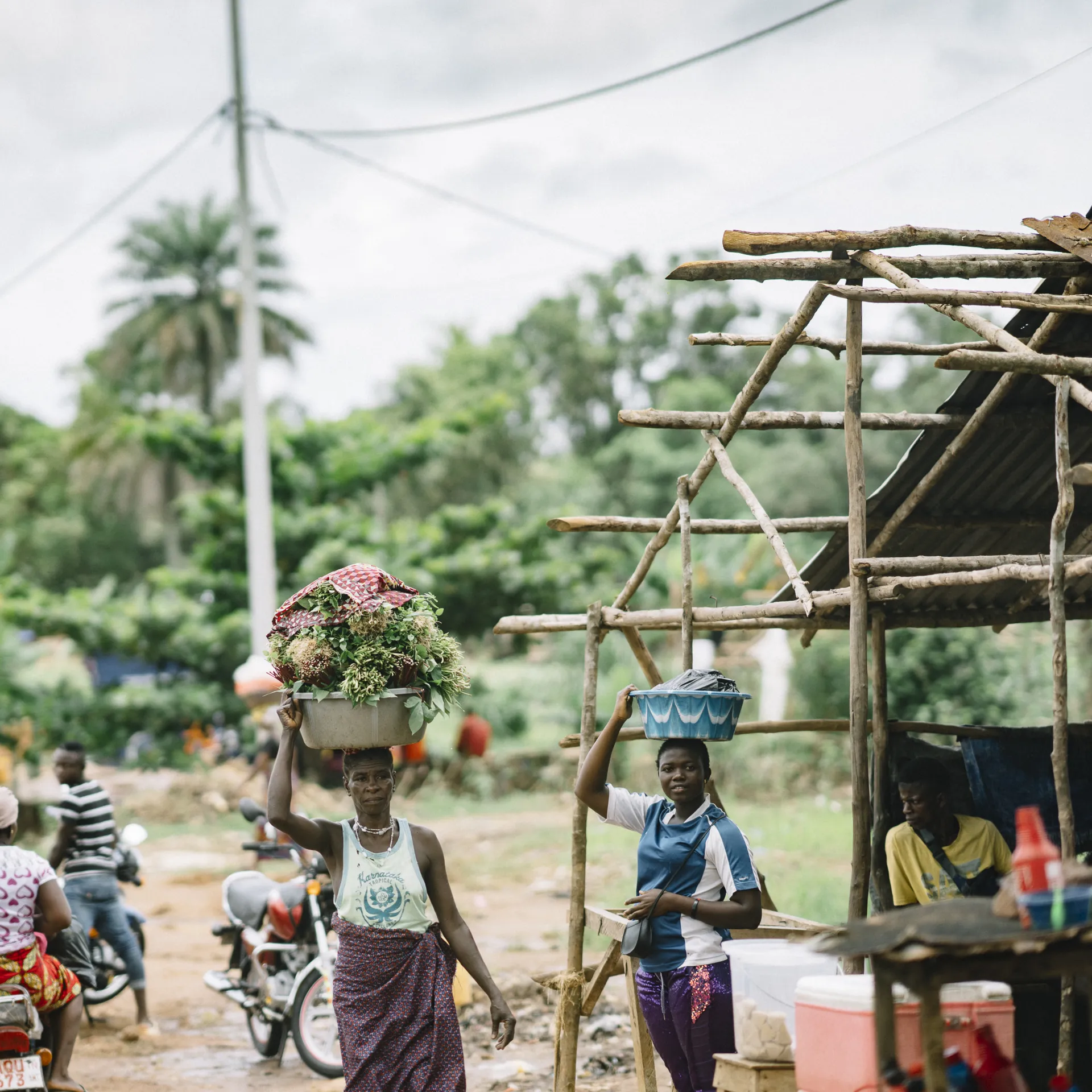 A woman balances a basket of flowers on her head while another carries goods. People and motorcycles are in the background, with lush greenery around.