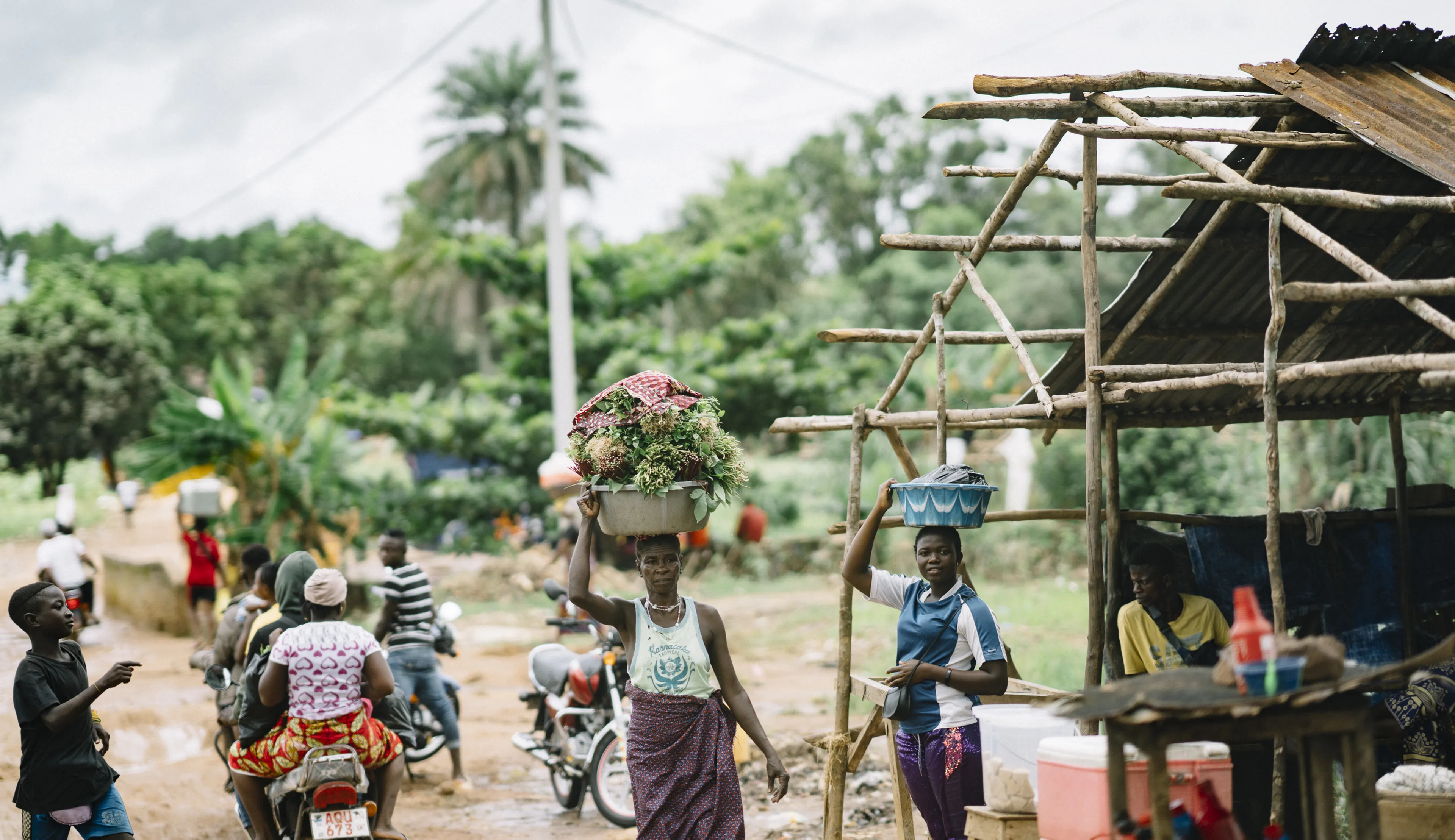 A woman balances a basket of flowers on her head while another carries goods. People and motorcycles are in the background, with lush greenery around.