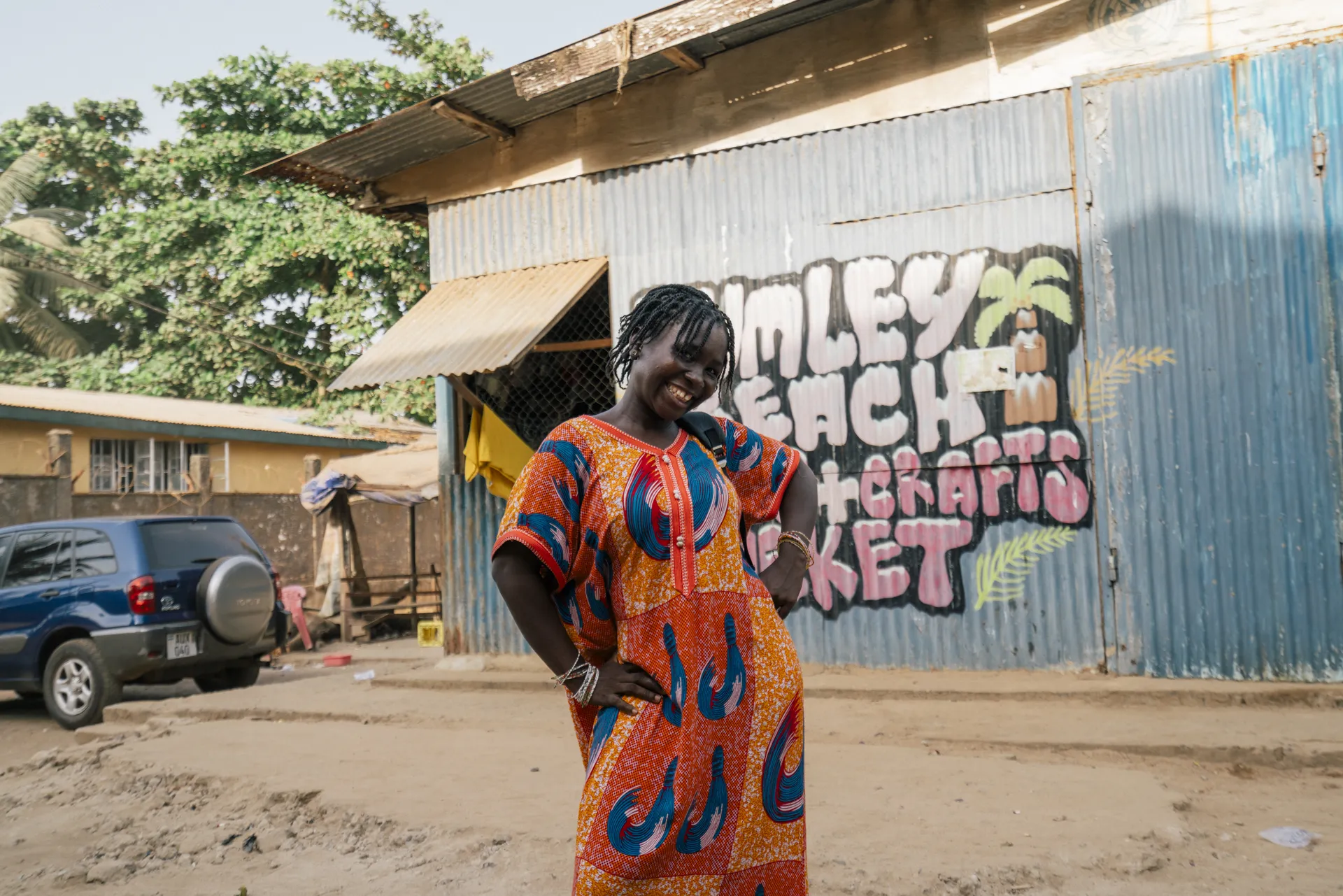 Woman in vibrant patterned dress smiles, standing in front of a corrugated metal building with colorful mural, trees in the background.