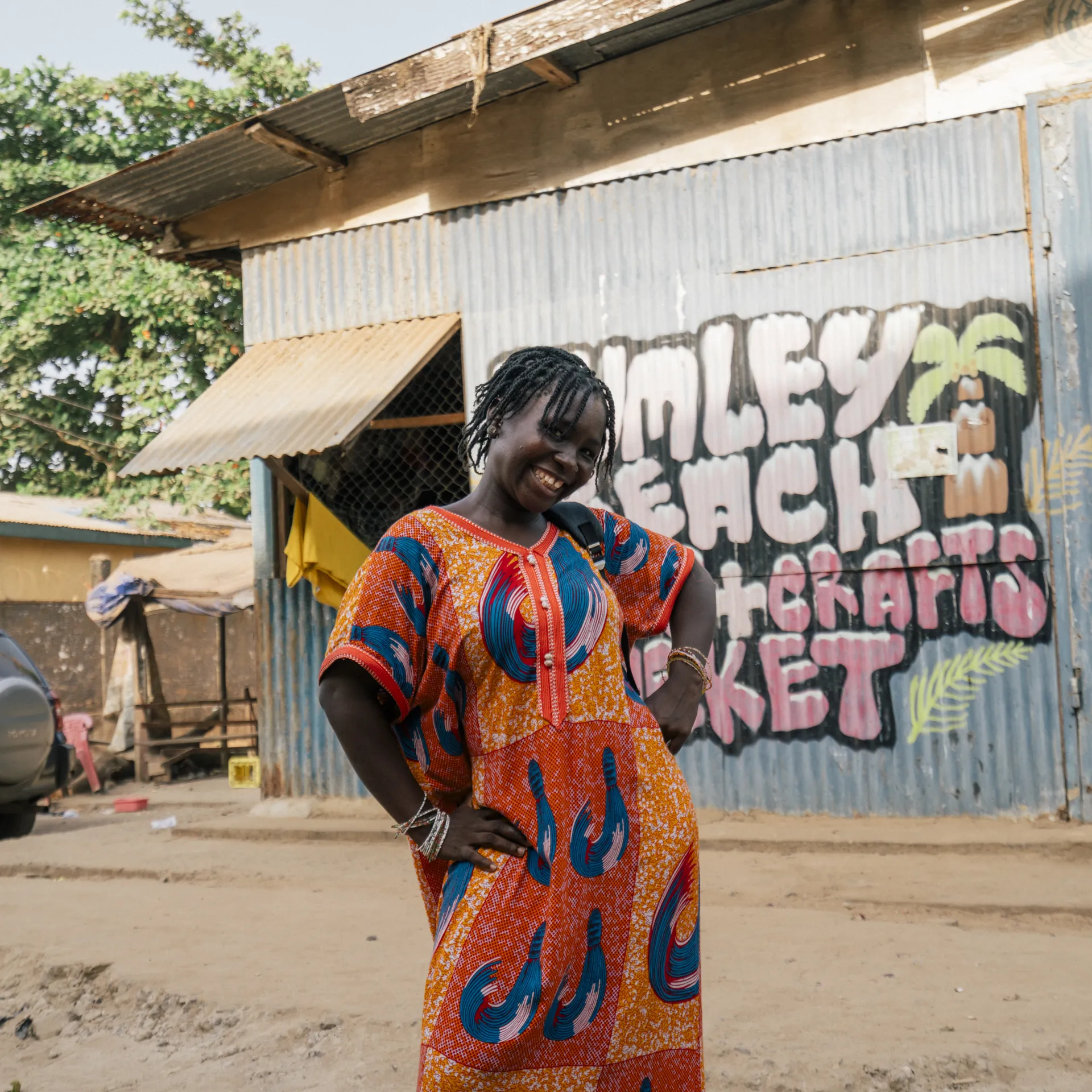 Woman in vibrant patterned dress smiles, standing in front of a corrugated metal building with colorful mural, trees in the background.