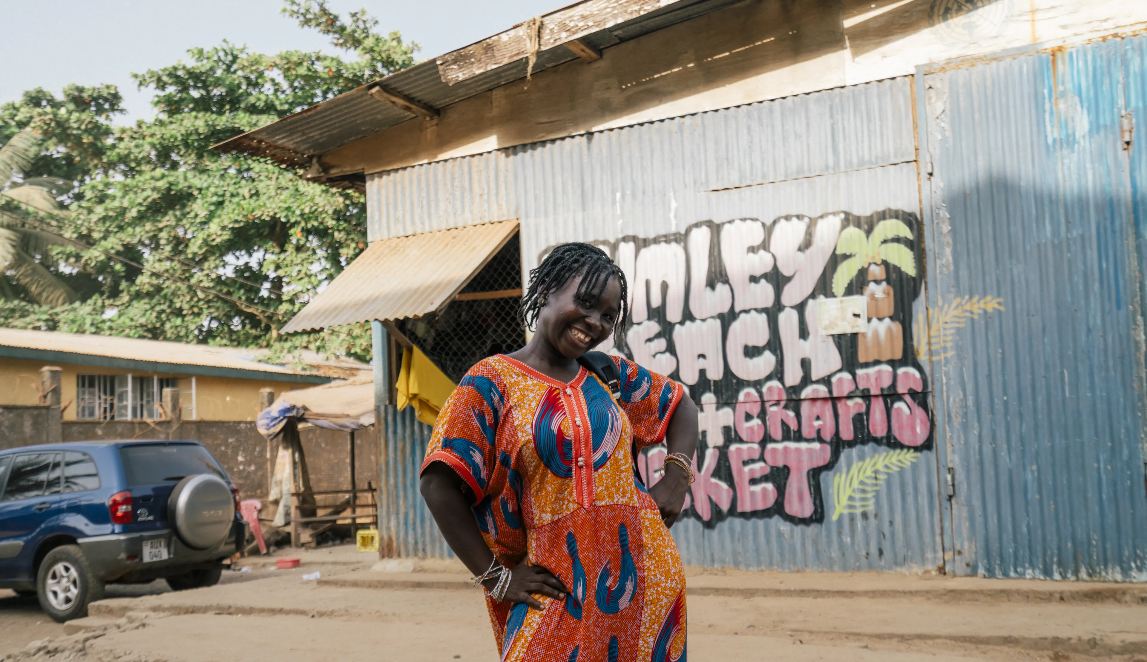 Woman in vibrant patterned dress smiles, standing in front of a corrugated metal building with colorful mural, trees in the background.