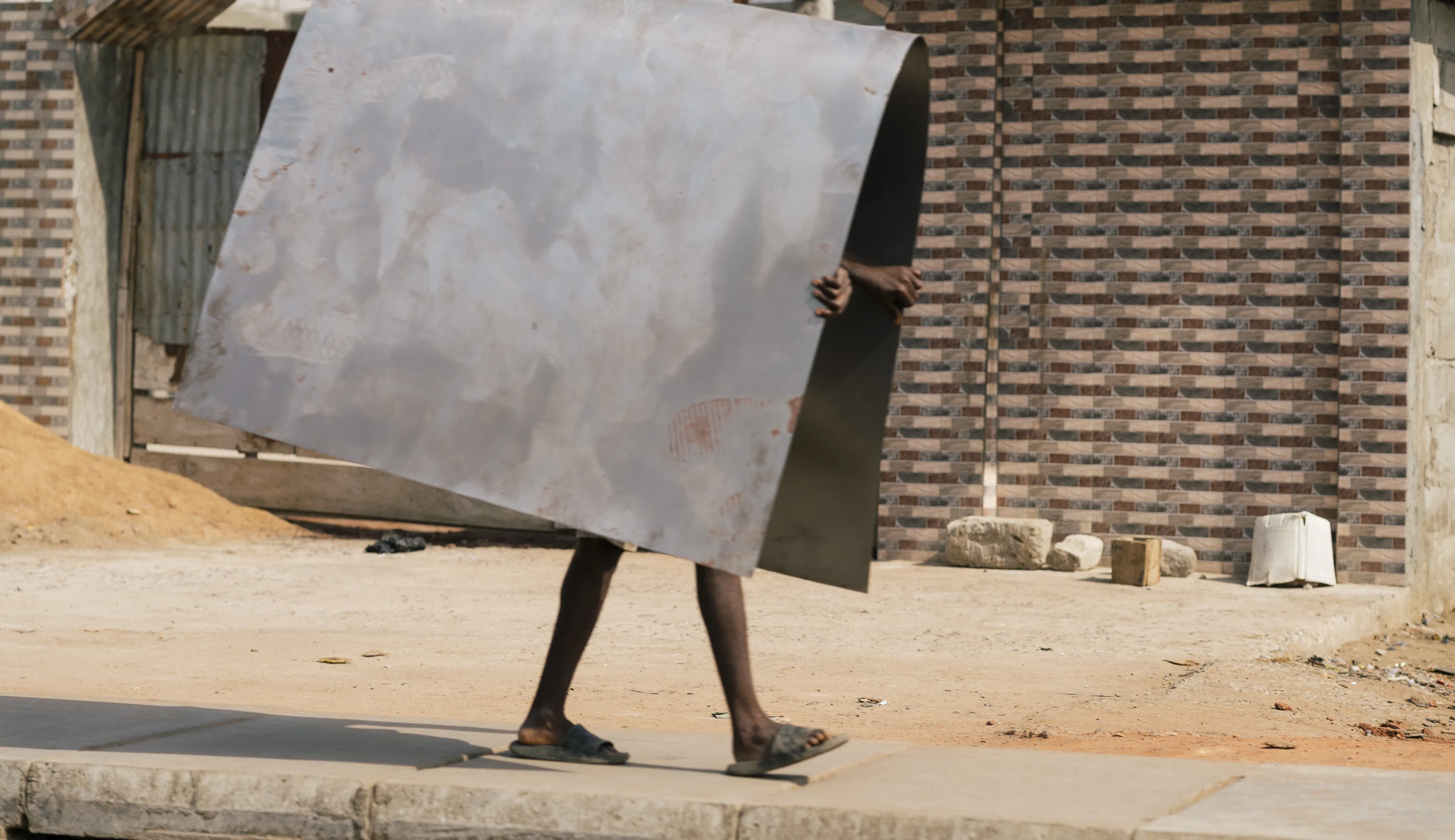 Person carrying a large sheet of metal on a sidewalk in front of a brick building, with only their legs visible.