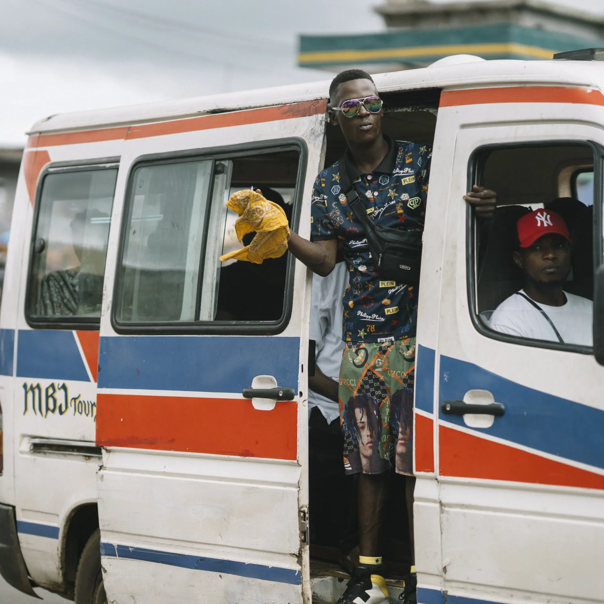 A person leans out of an open minibus door holding a plastic bag, while another sits inside. The minibus is white with red and blue stripes.