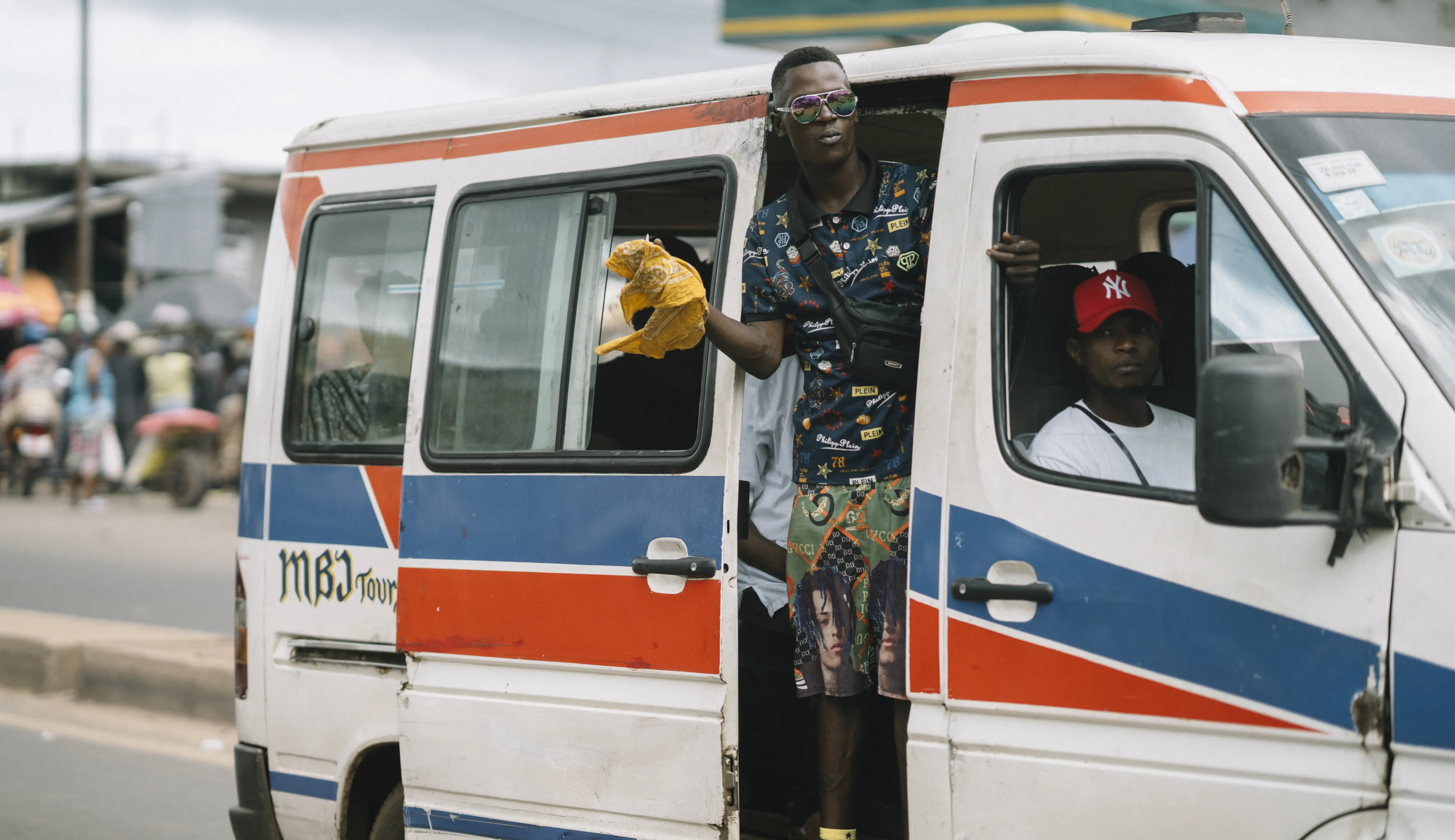A person leans out of an open minibus door holding a plastic bag, while another sits inside. The minibus is white with red and blue stripes.