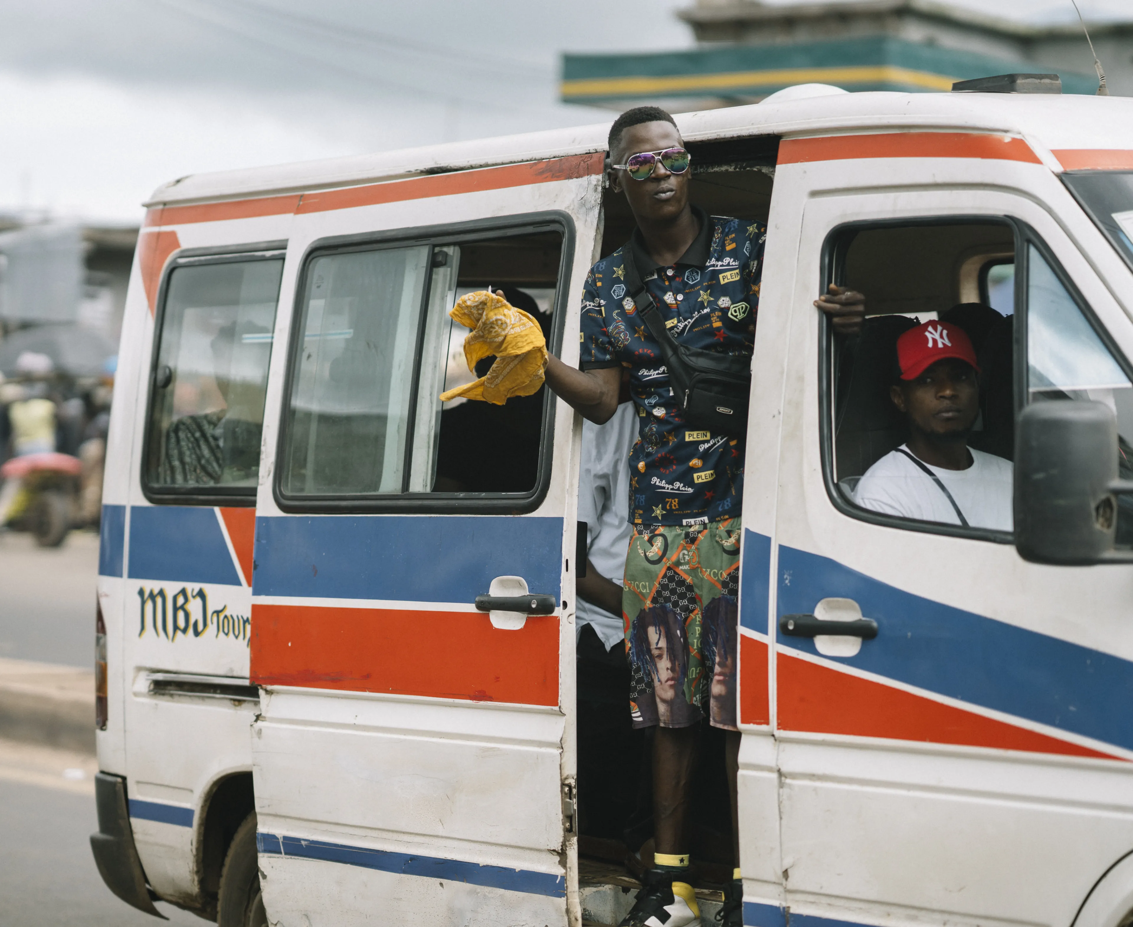 A person leans out of an open minibus door holding a plastic bag, while another sits inside. The minibus is white with red and blue stripes.