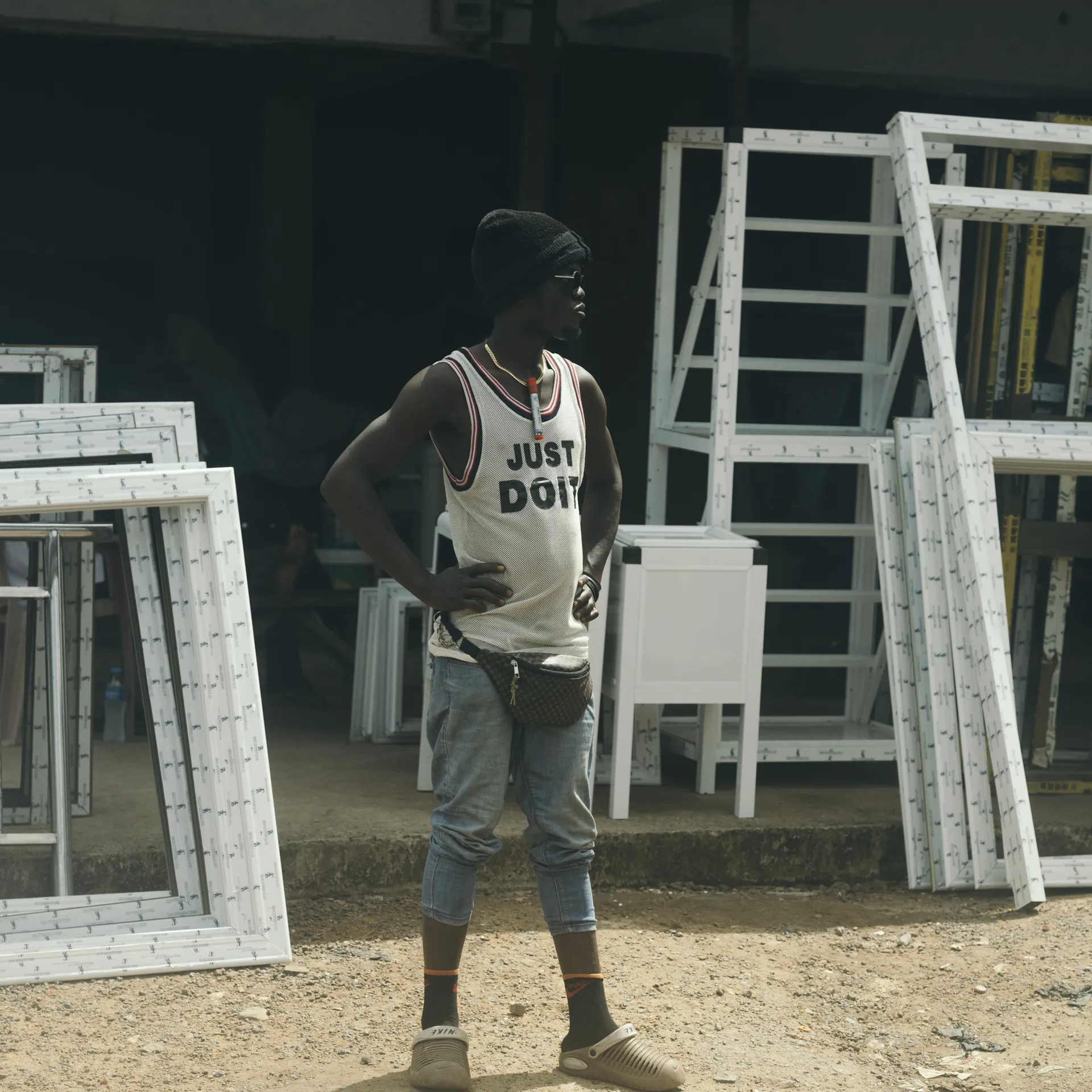 Person standing outdoors among stacked window frames, wearing a white tank top and a beanie, with hands on hips.
