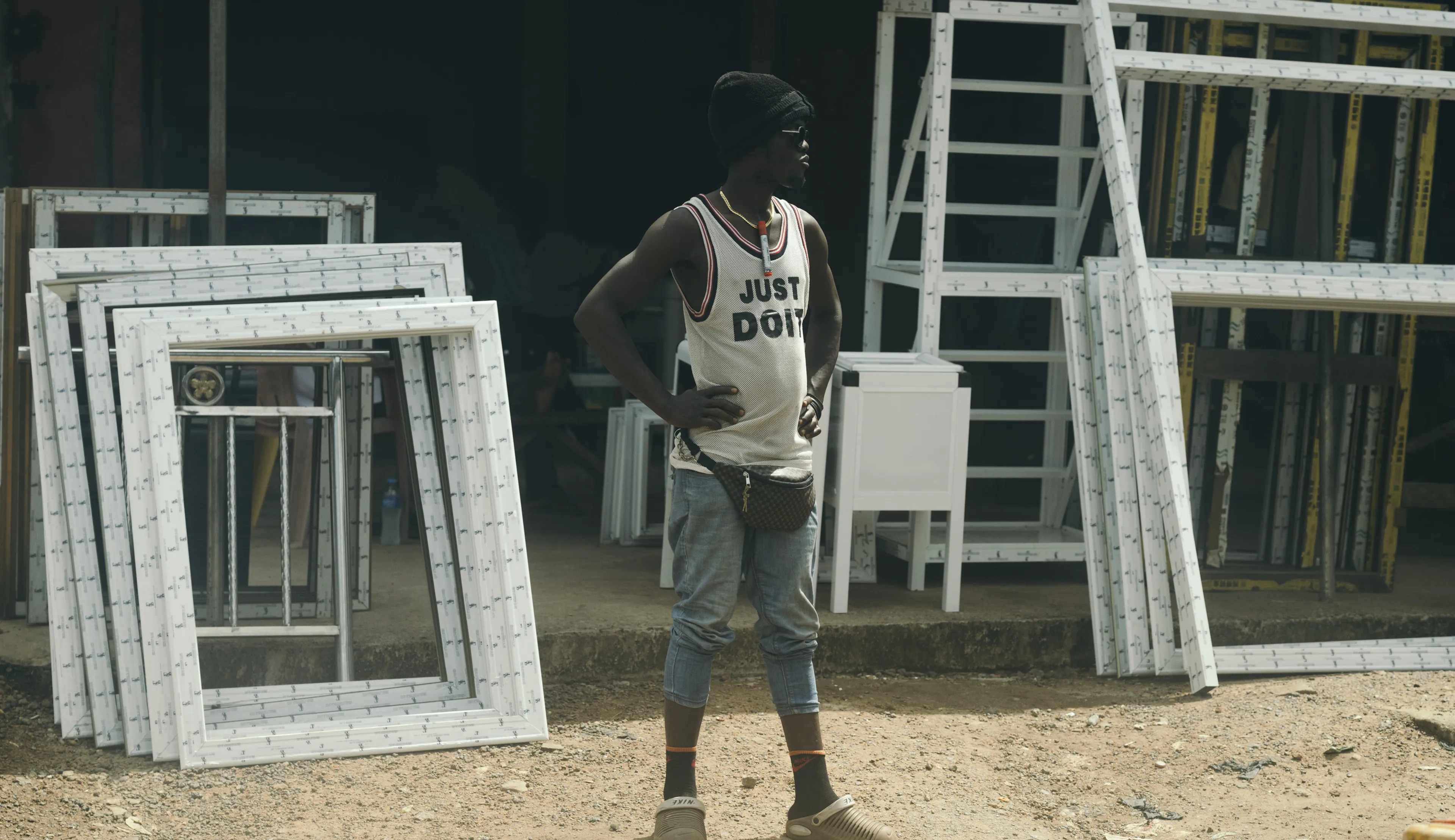 Person standing outdoors among stacked window frames, wearing a white tank top and a beanie, with hands on hips.