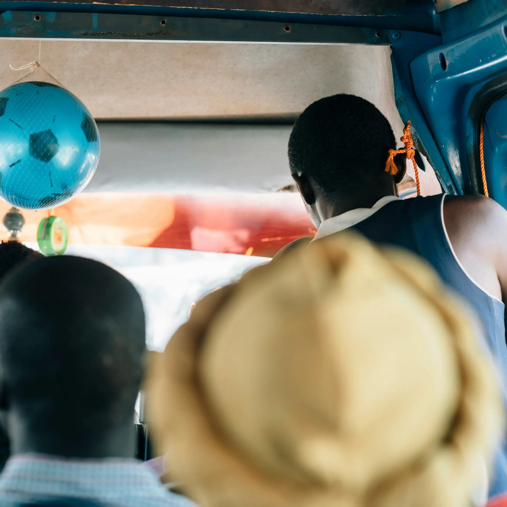 People sitting in a crowded bus, with a blue football hanging from the ceiling. A person stands near the door, and the interior is vibrant.