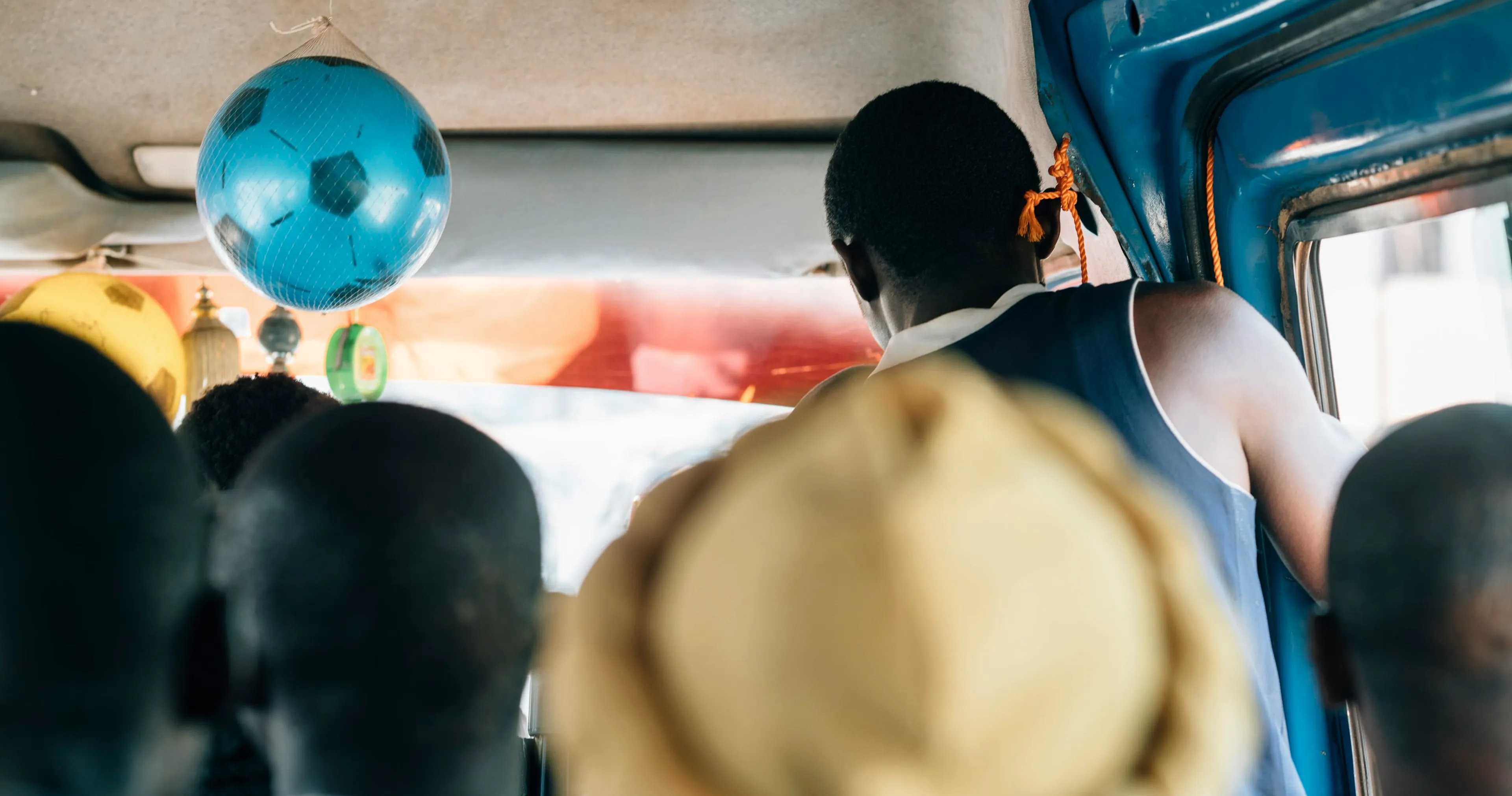 People sitting in a crowded bus, with a blue football hanging from the ceiling. A person stands near the door, and the interior is vibrant.