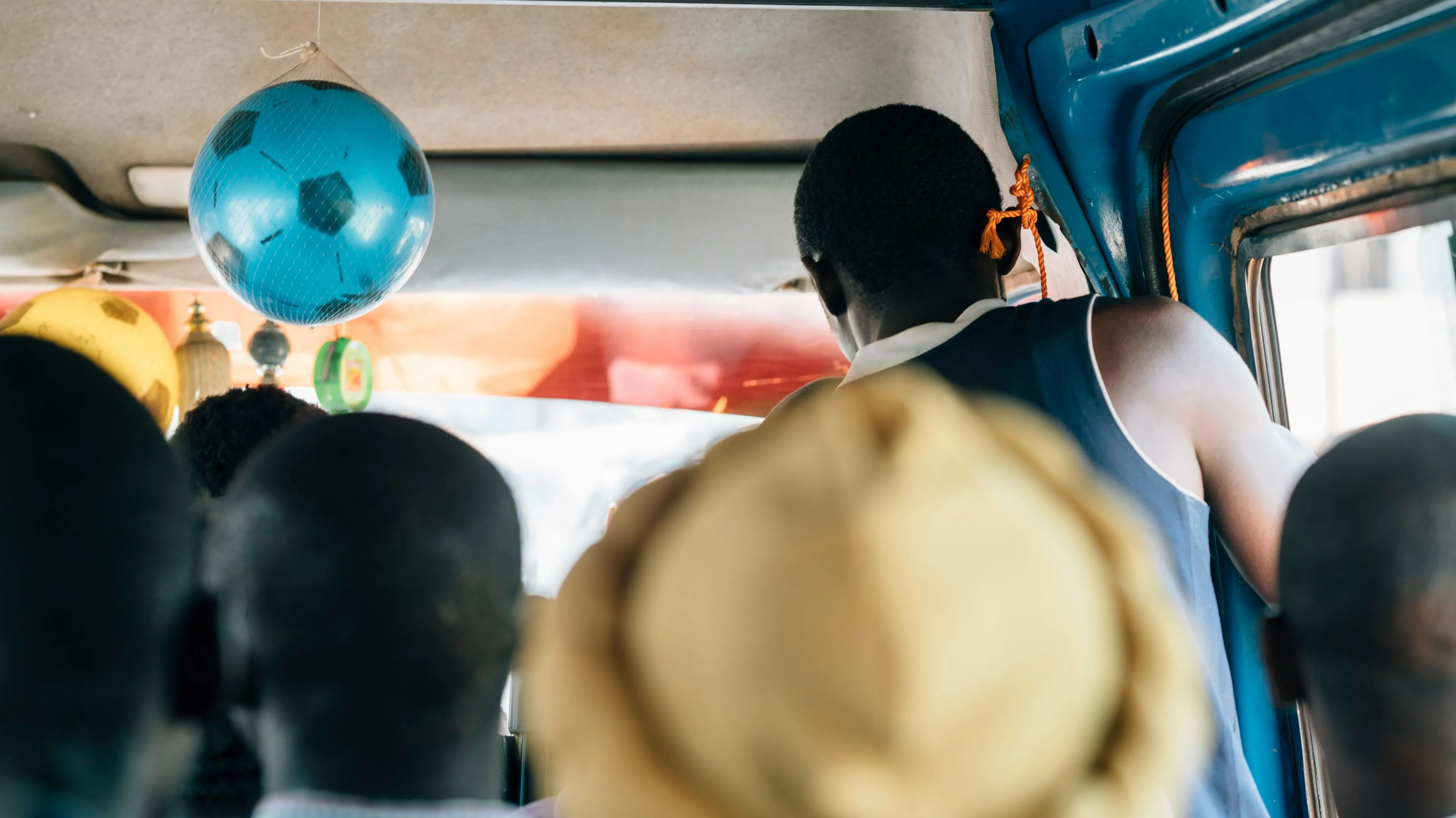 People sitting in a crowded bus, with a blue football hanging from the ceiling. A person stands near the door, and the interior is vibrant.