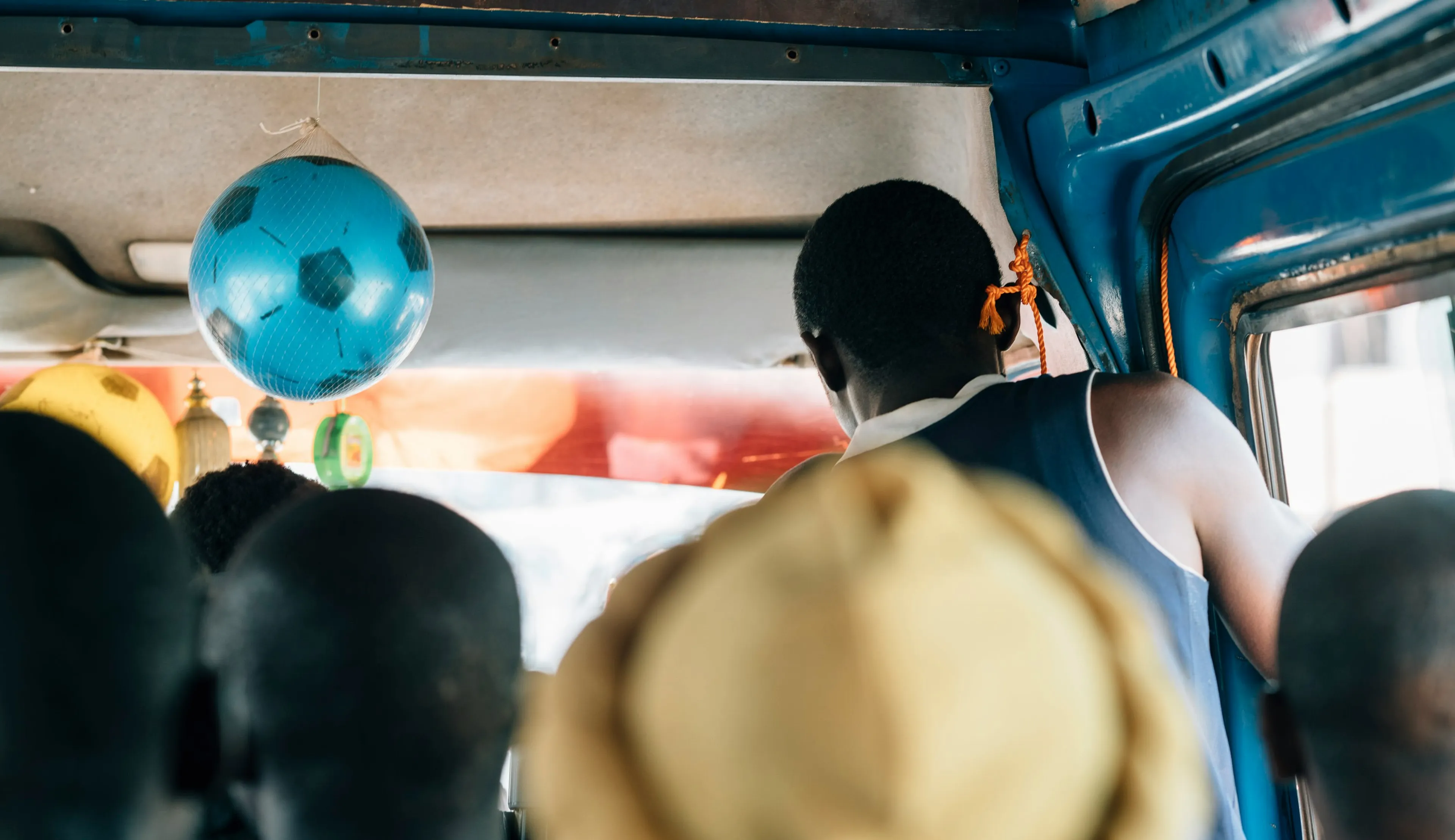 People sitting in a crowded bus, with a blue football hanging from the ceiling. A person stands near the door, and the interior is vibrant.