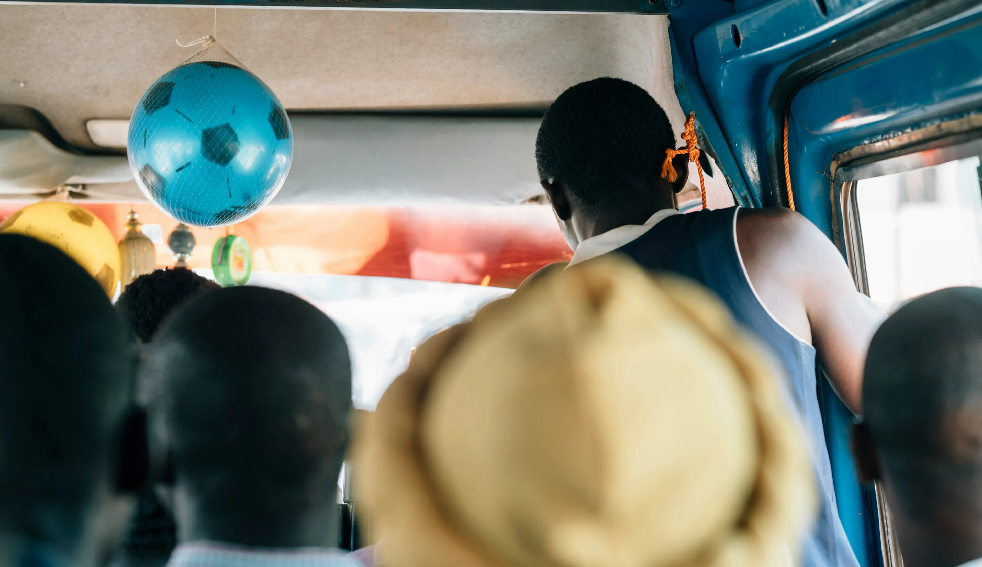 People sitting in a crowded bus, with a blue football hanging from the ceiling. A person stands near the door, and the interior is vibrant.