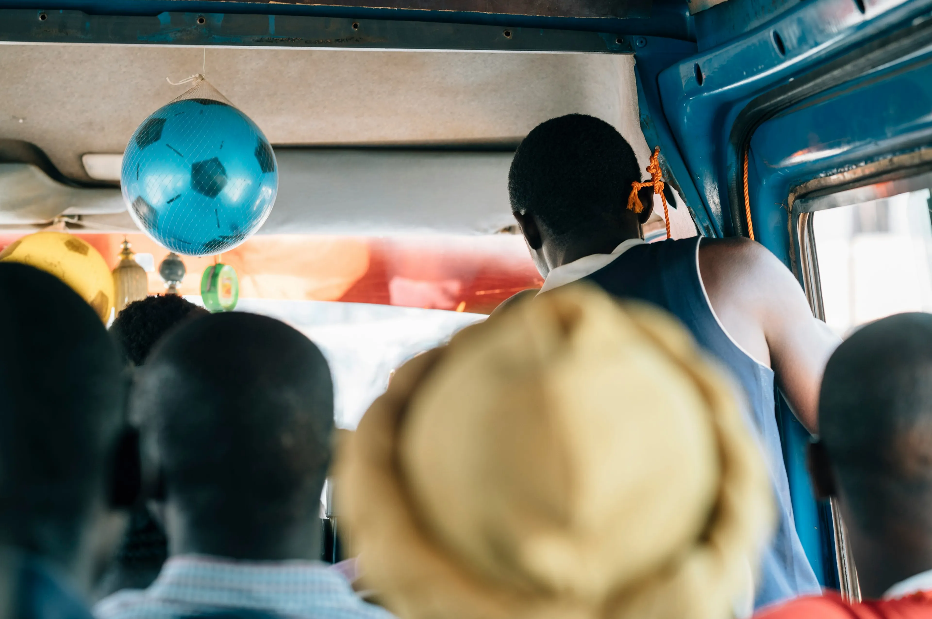 People sitting in a crowded bus, with a blue football hanging from the ceiling. A person stands near the door, and the interior is vibrant.
