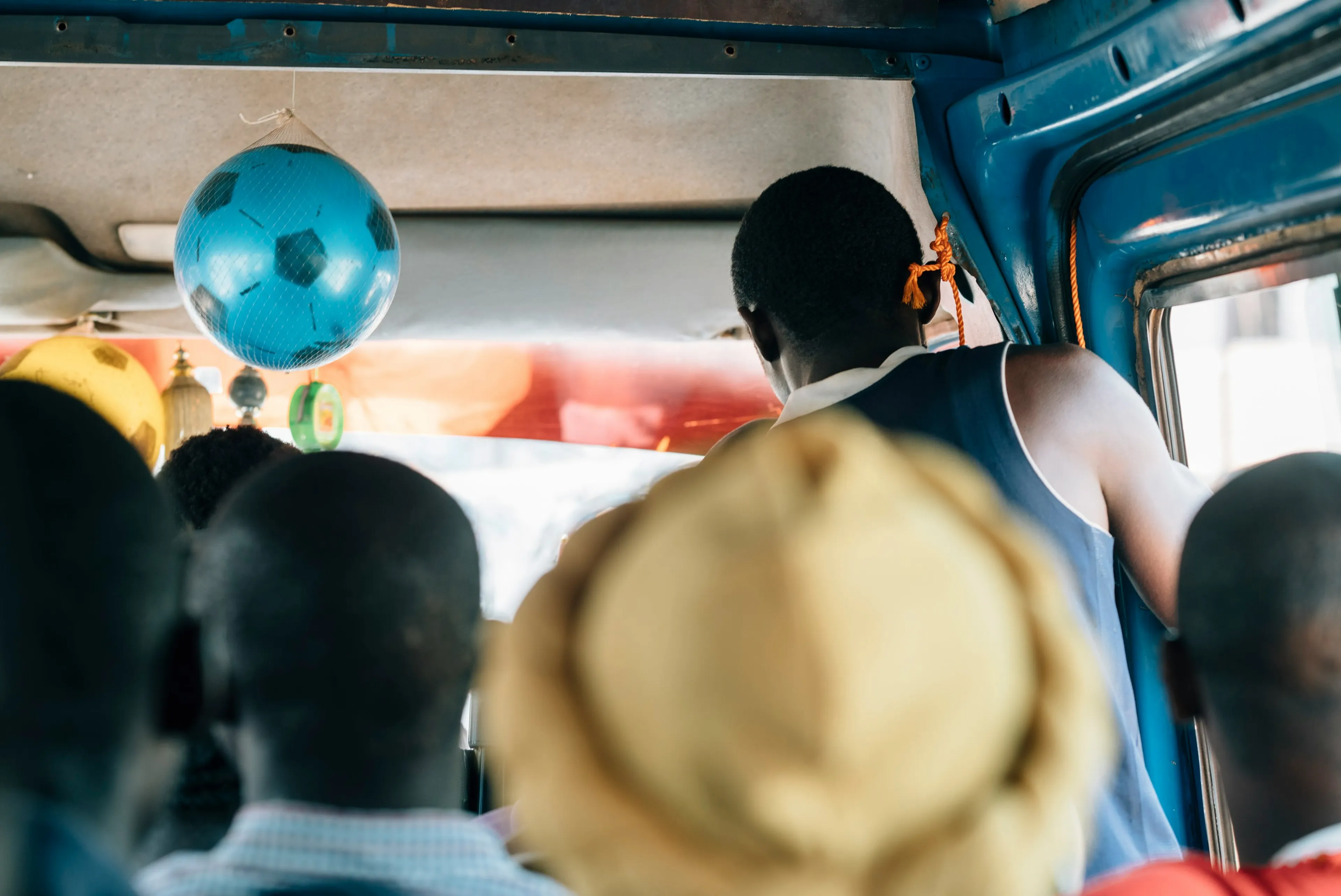 People sitting in a crowded bus, with a blue football hanging from the ceiling. A person stands near the door, and the interior is vibrant.