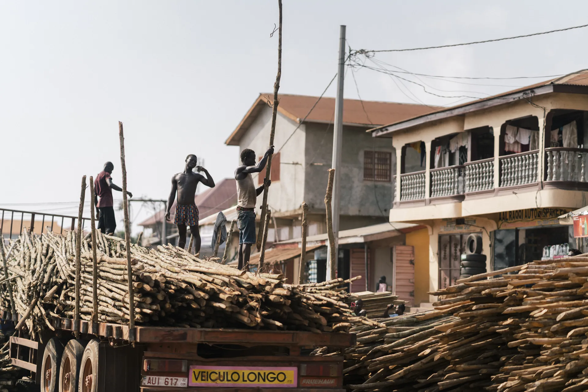 Workers loading long wooden logs onto a truck in a busy street with buildings and power lines in the background.
