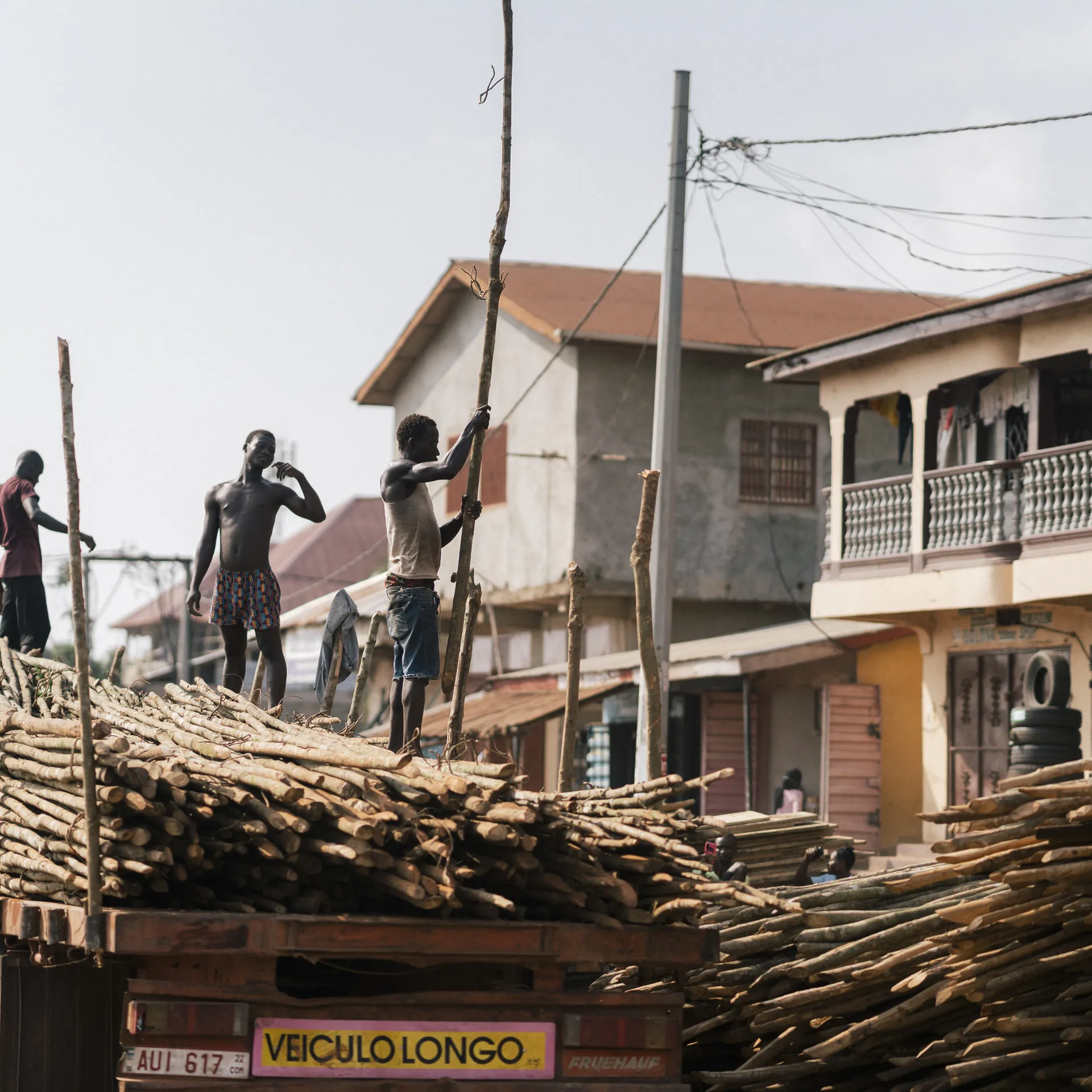 Workers loading long wooden logs onto a truck in a busy street with buildings and power lines in the background.