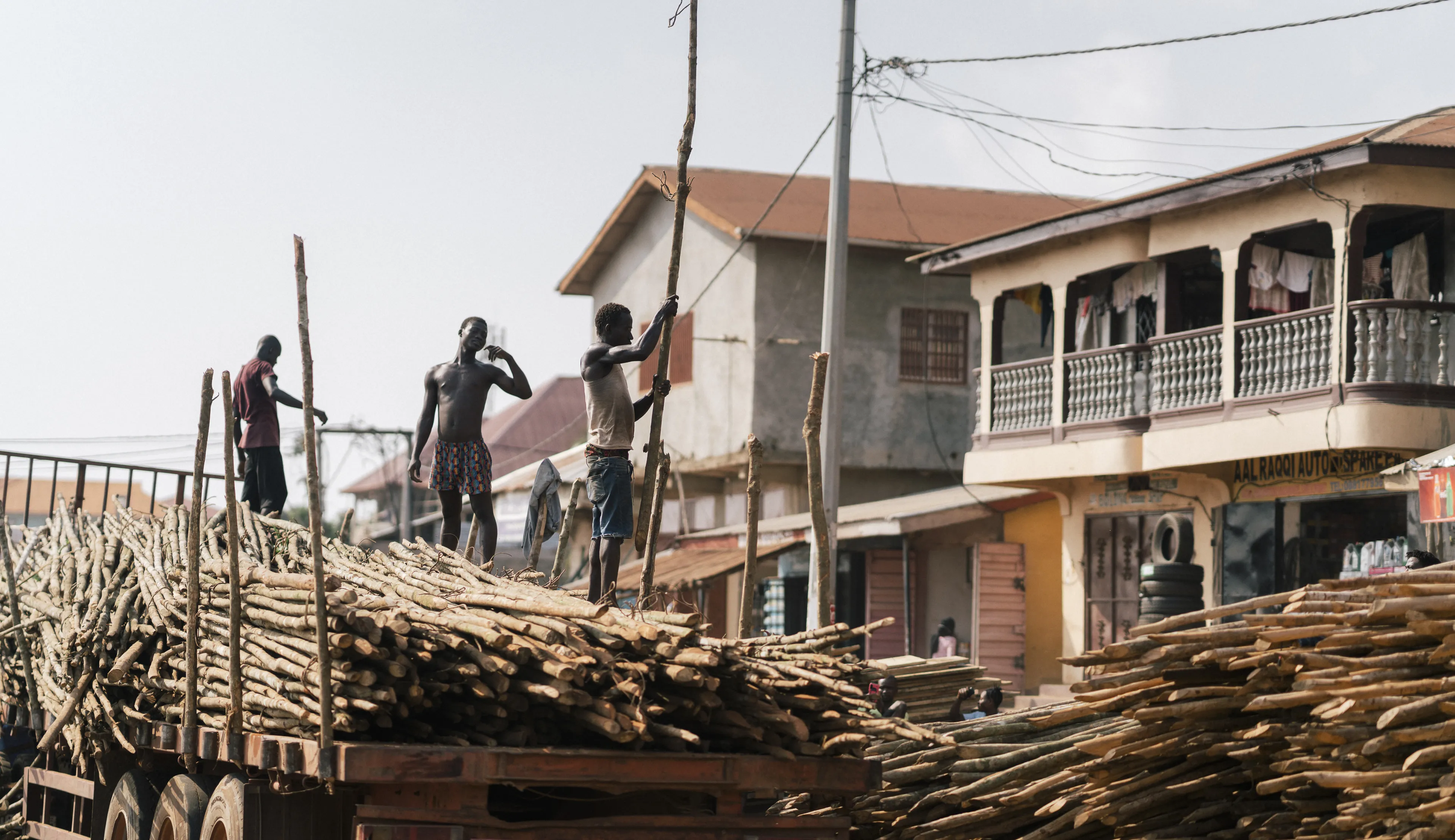 Workers loading long wooden logs onto a truck in a busy street with buildings and power lines in the background.