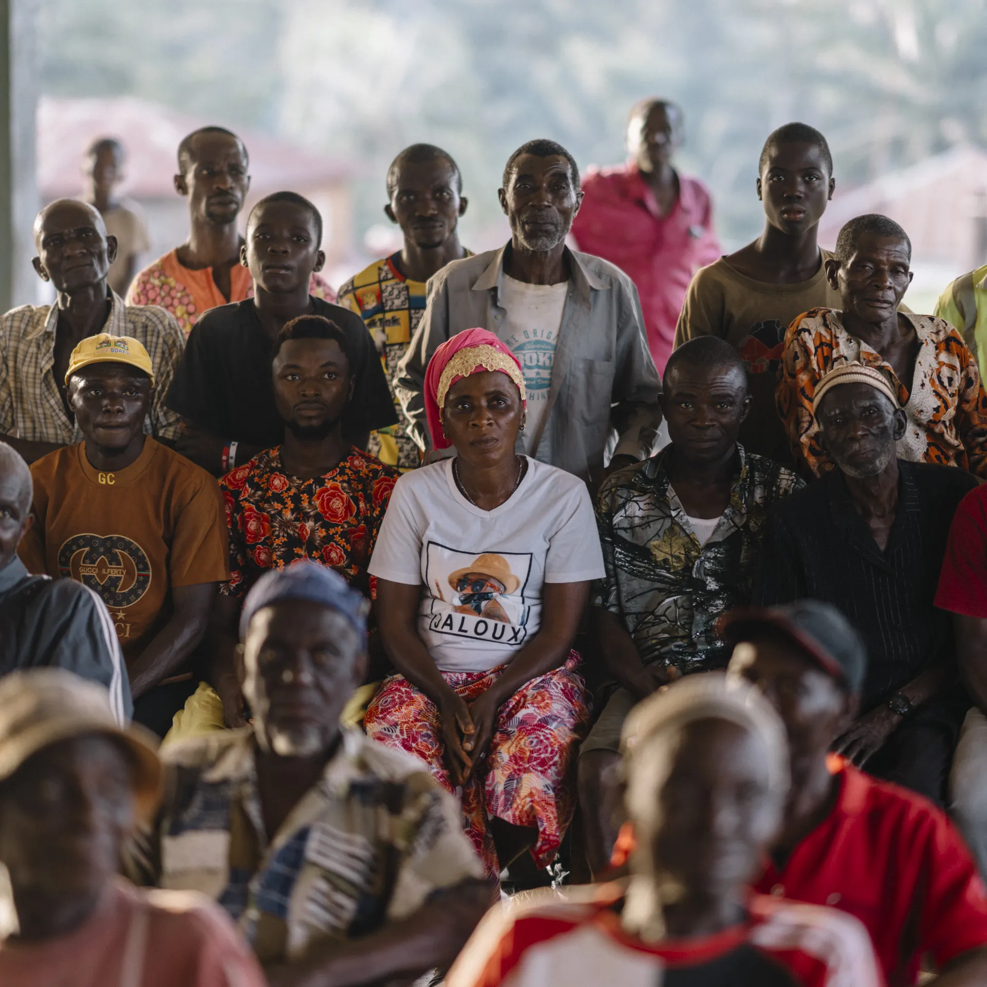 Group of African community members seated at a covered outdoor meeting, facing the camera with attentive expressions.