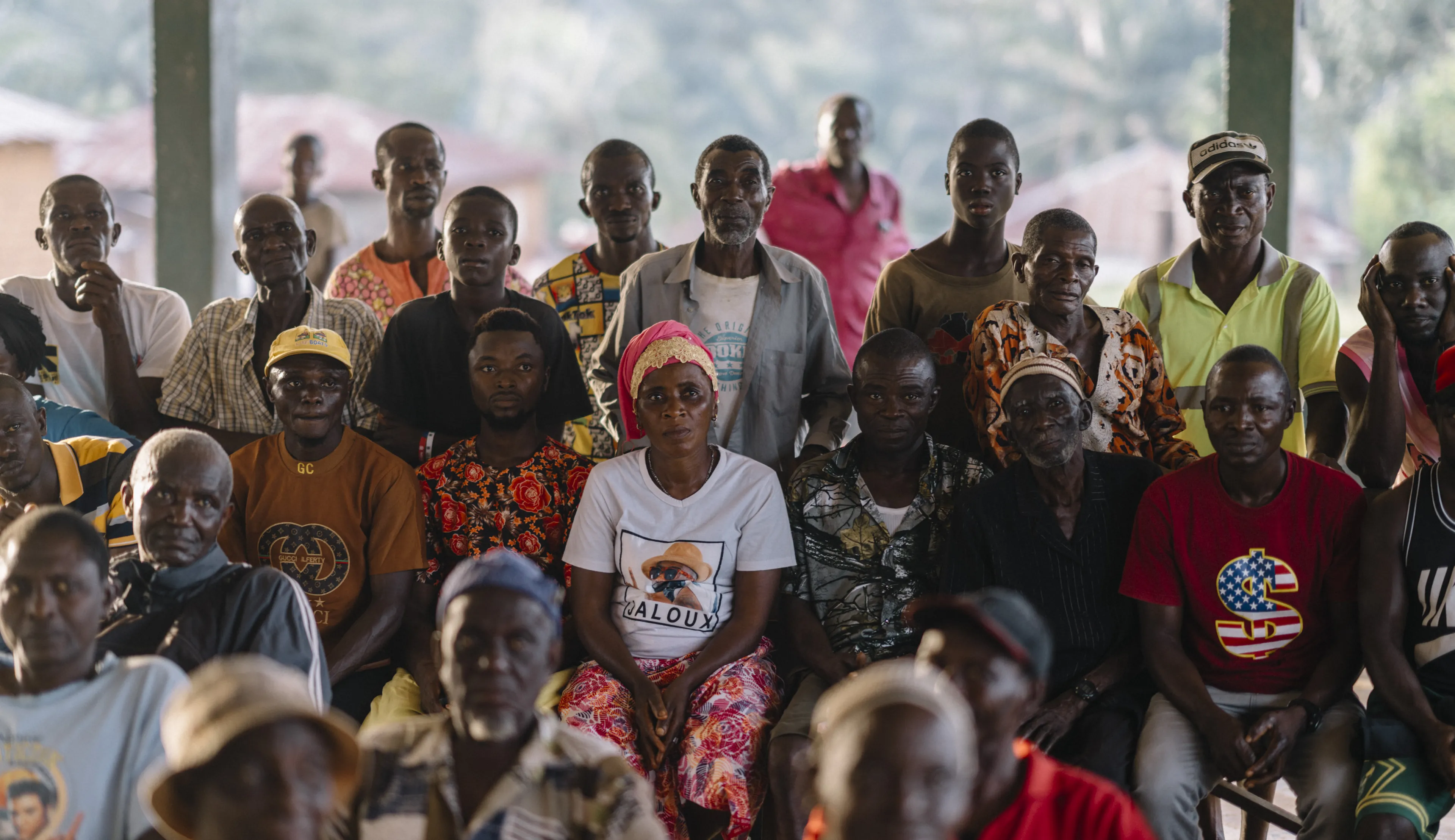 Group of African community members seated at a covered outdoor meeting, facing the camera with attentive expressions.