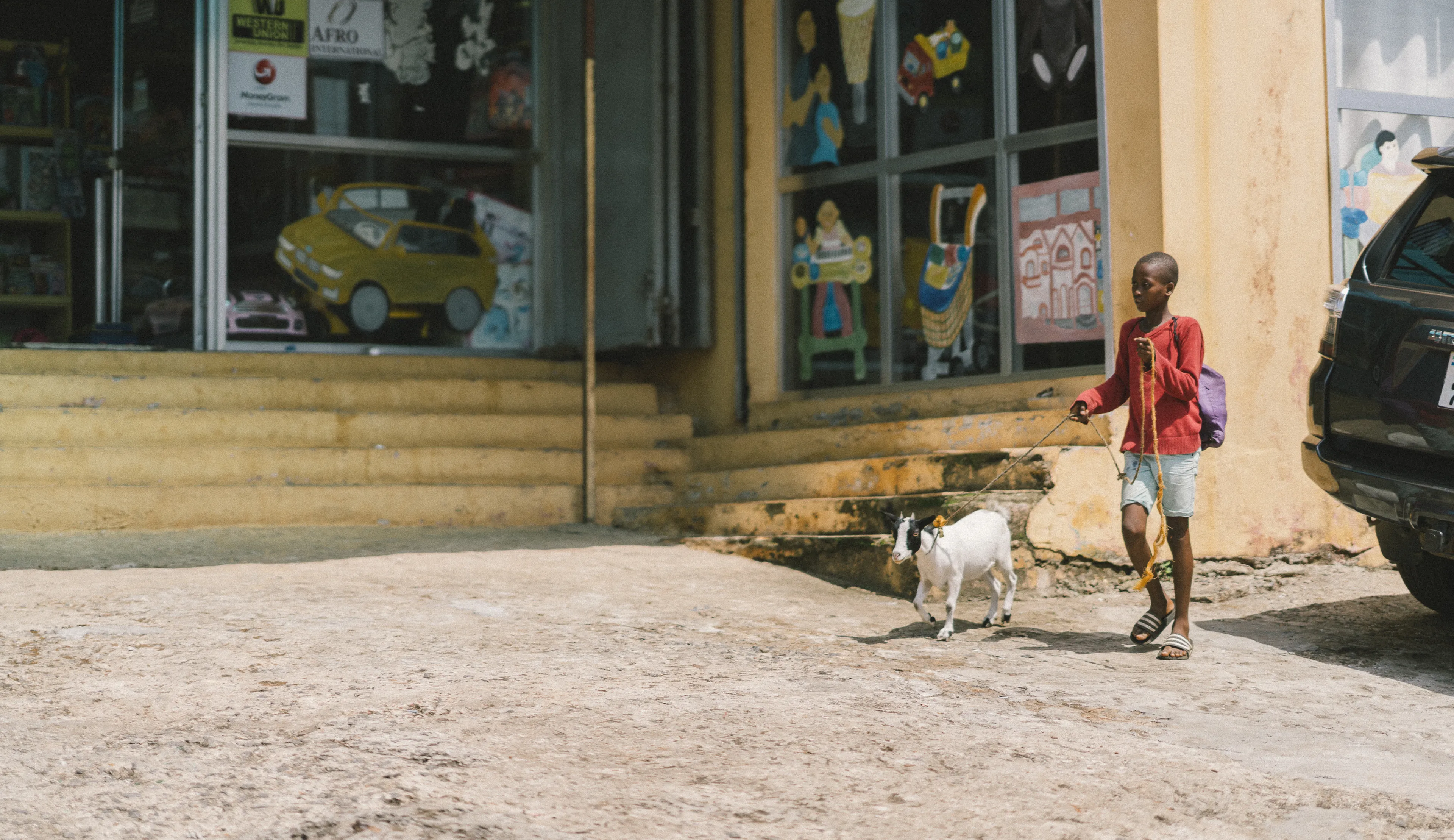 A child in a red shirt walks a small goat on a leash near a storefront with colorful signs and a parked car.