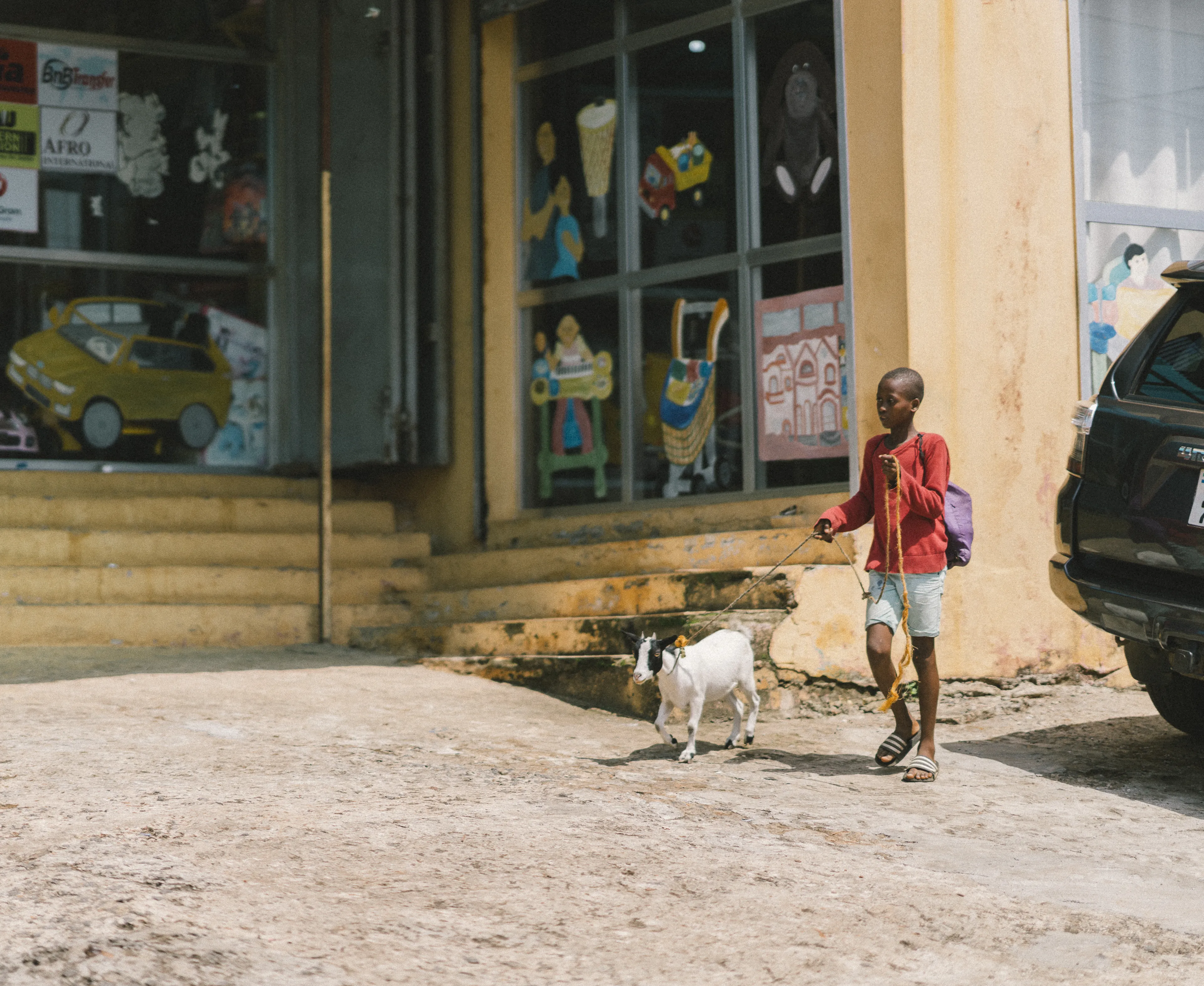 A child in a red shirt walks a small goat on a leash near a storefront with colorful signs and a parked car.