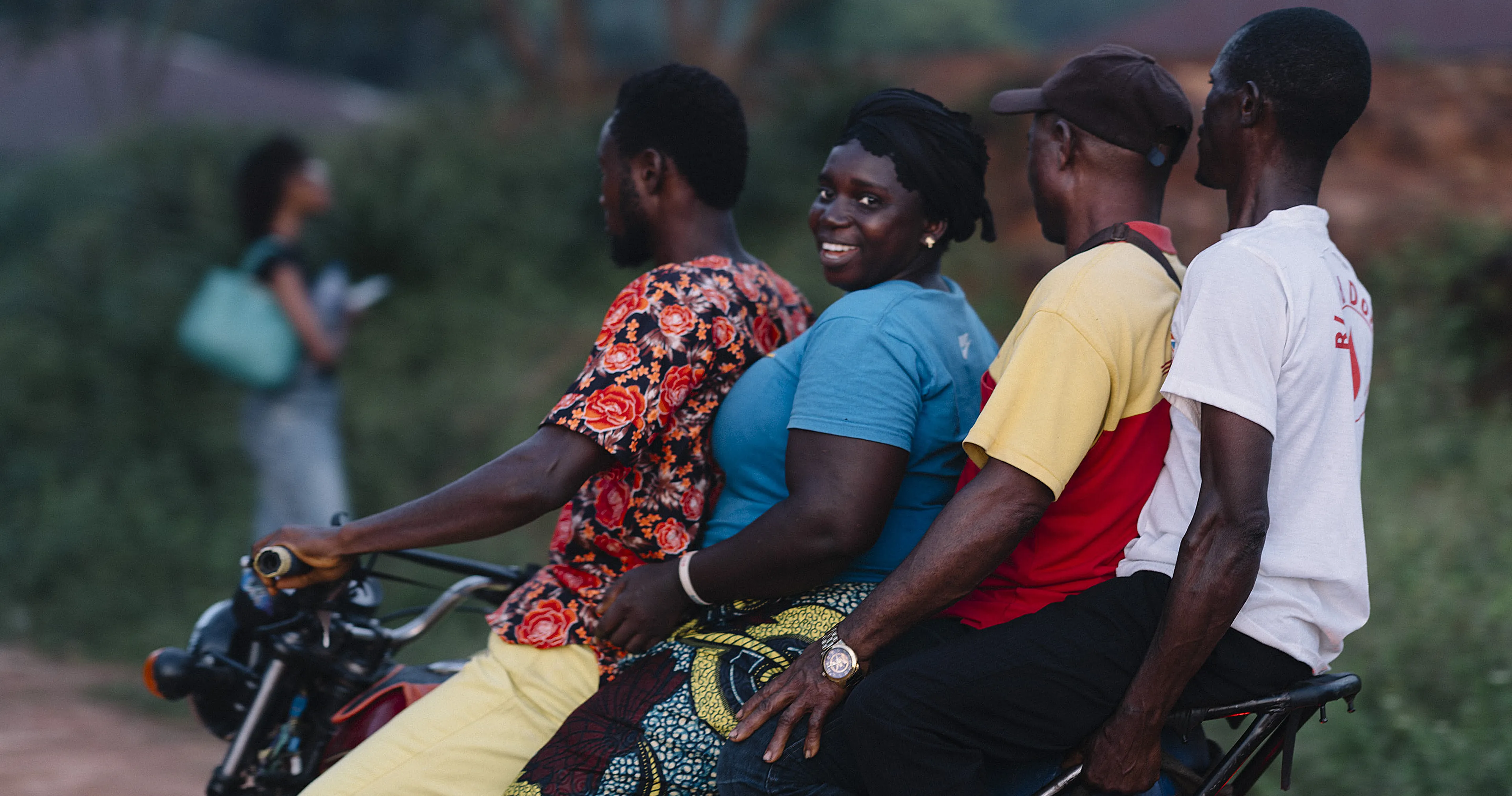 Four people ride a motorcycle together on a rural road; the woman in blue smiles at the camera.