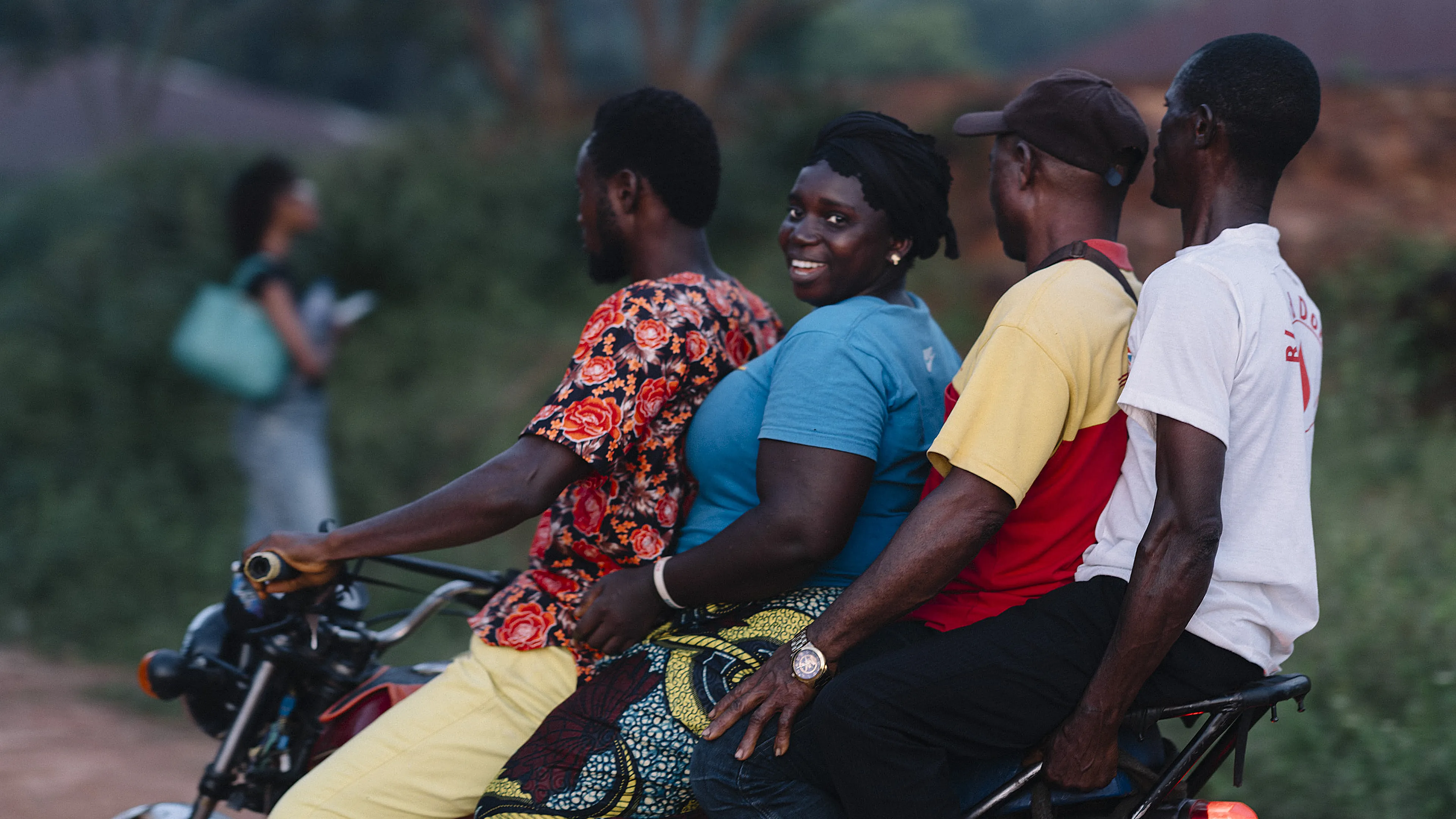 Four people ride a motorcycle together on a rural road; the woman in blue smiles at the camera.