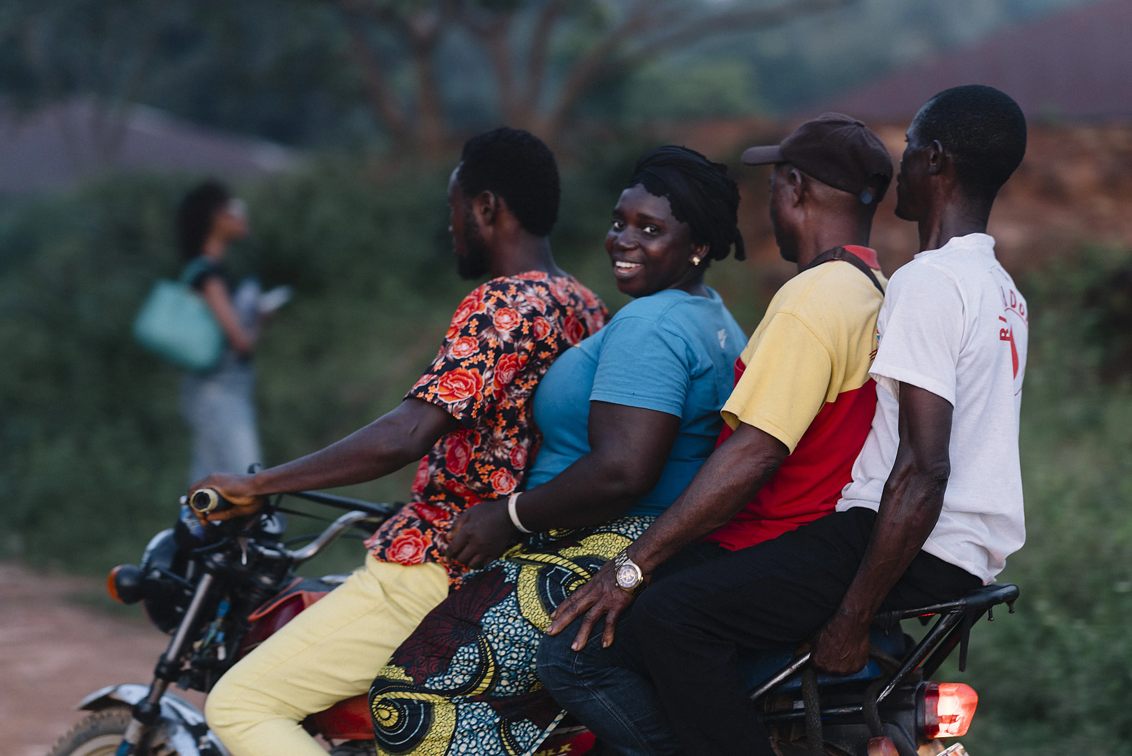 Four people ride a motorcycle together on a rural road; the woman in blue smiles at the camera.