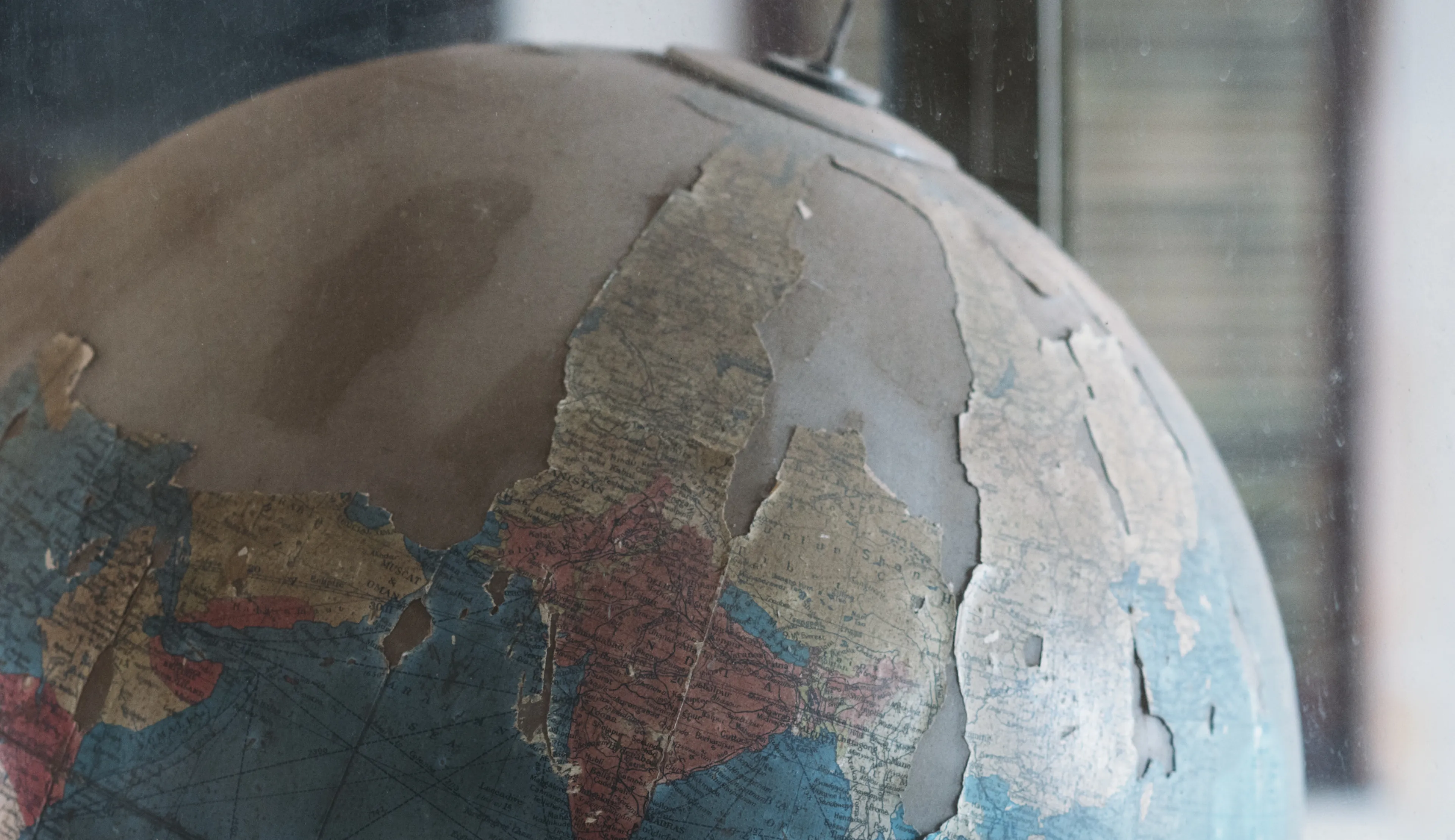 Close-up of a worn,  globe with peeling paper, showing faded blue seas and tan continents, near a blurred background.