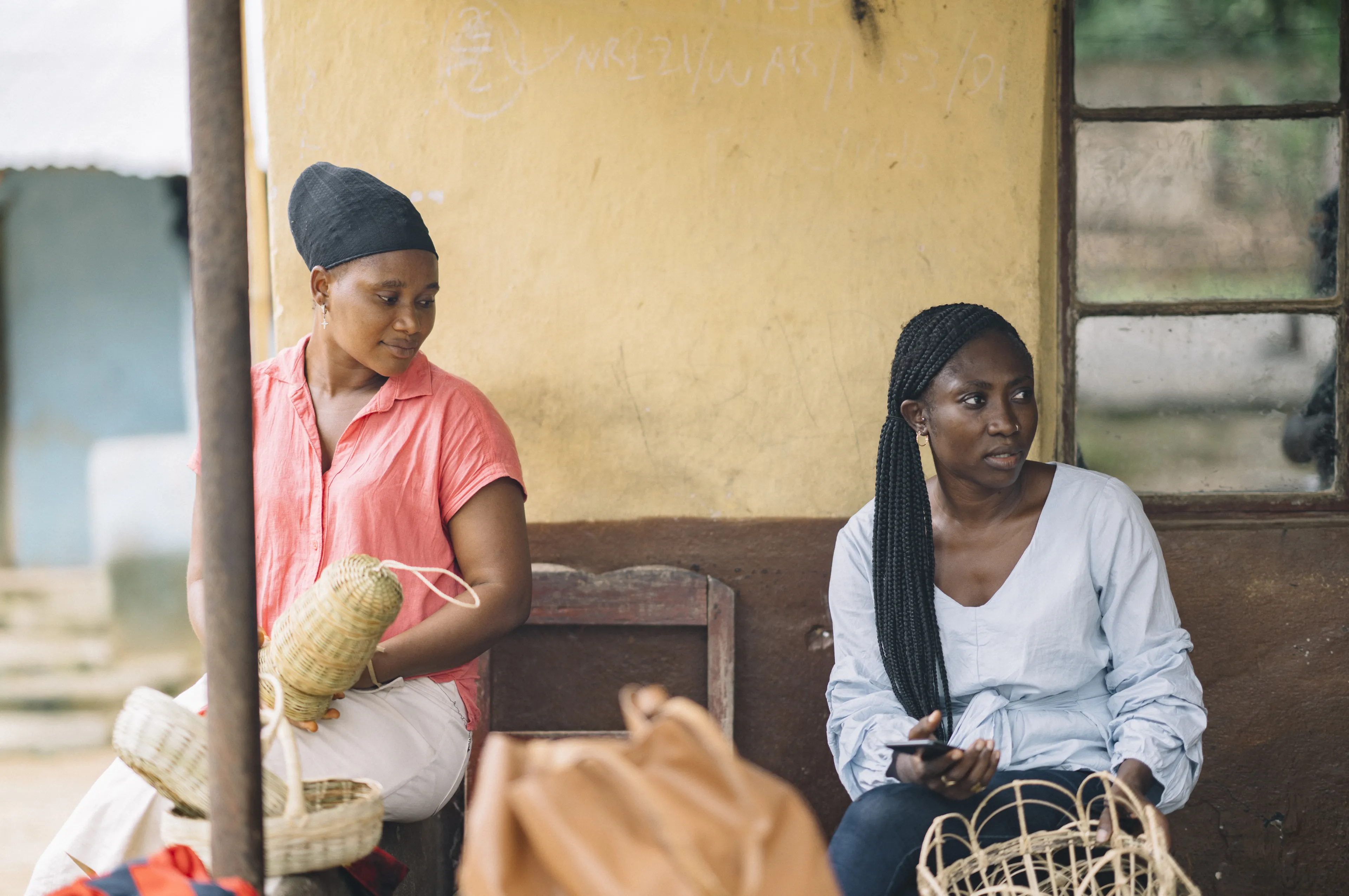 Two women sitting outside, weaving baskets. One holds a basket and the other looks aside, holding a phone. A yellow wall is in the background.