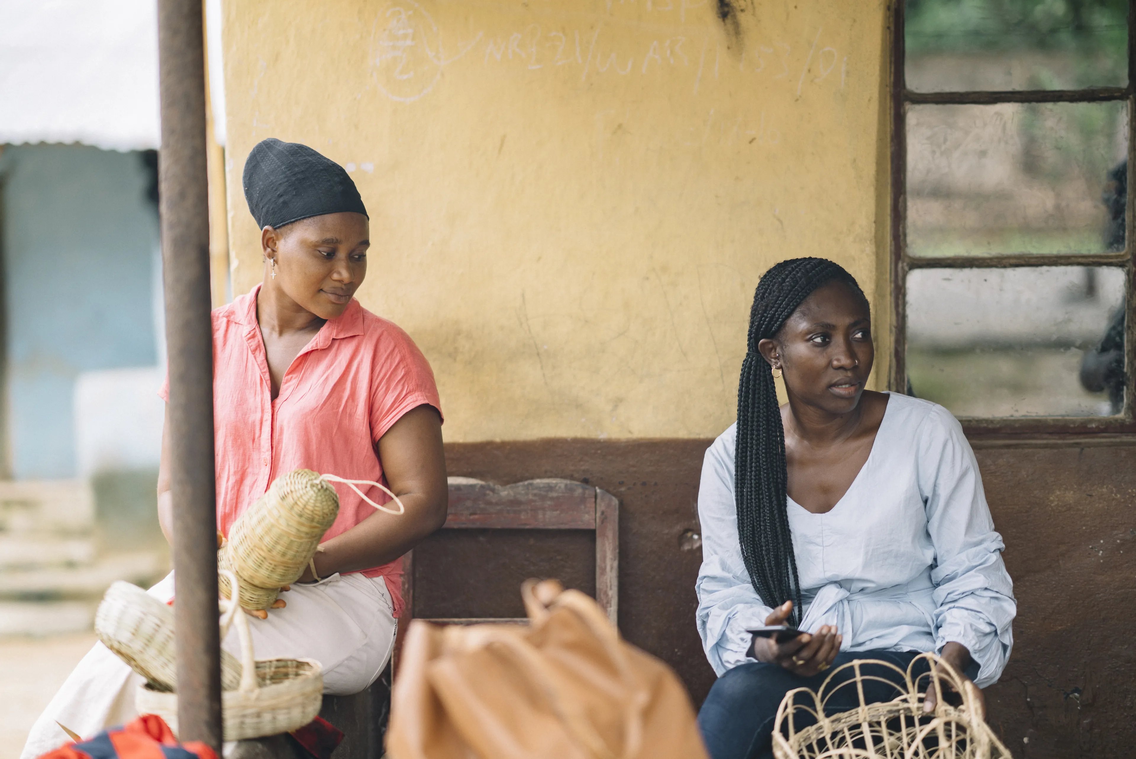 Two women sitting outside, weaving baskets. One holds a basket and the other looks aside, holding a phone. A yellow wall is in the background.