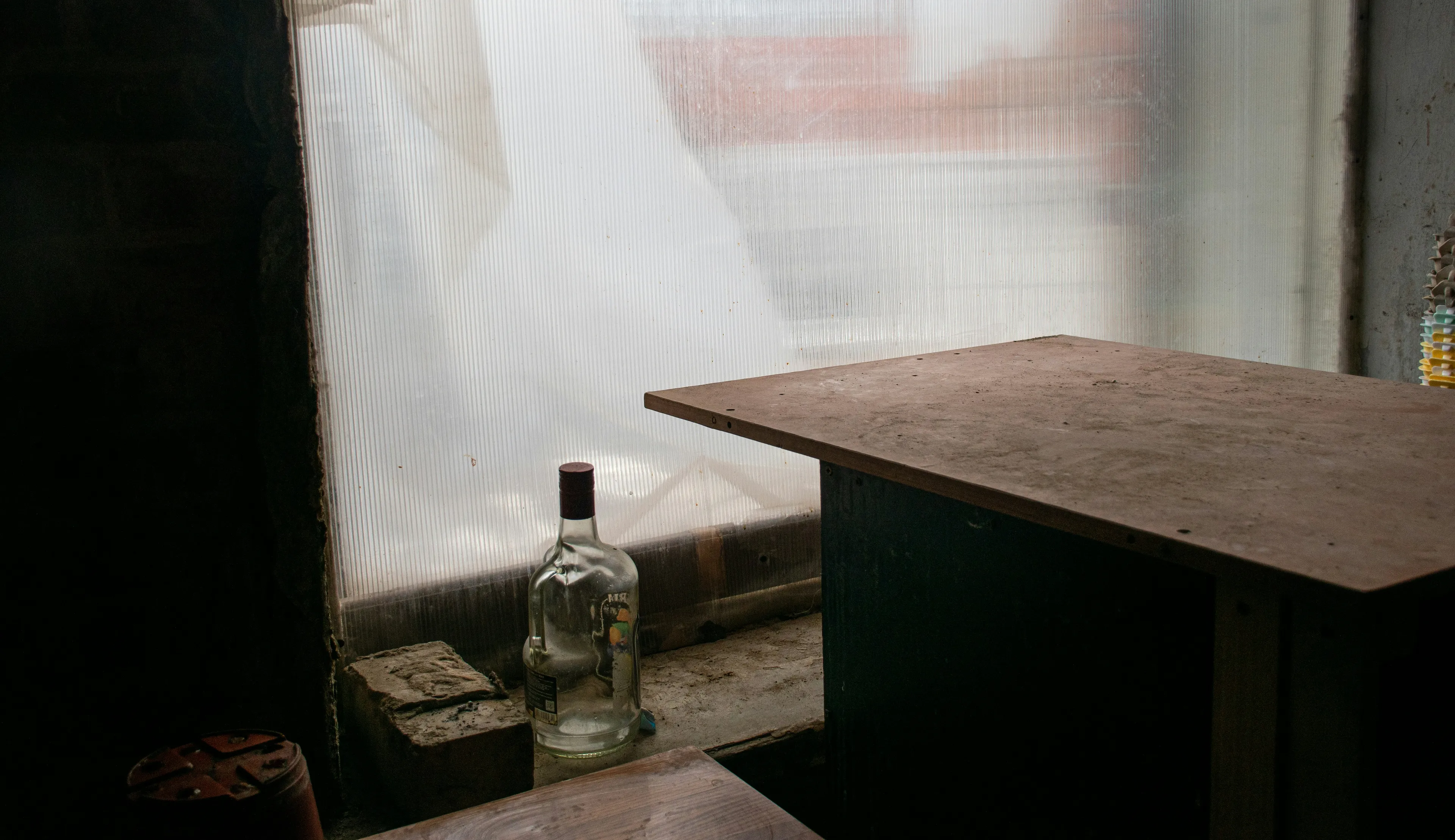 A dimly lit room with a frosted window, a wooden table, and a glass bottle on the ledge, creating a rustic, abandoned atmosphere.