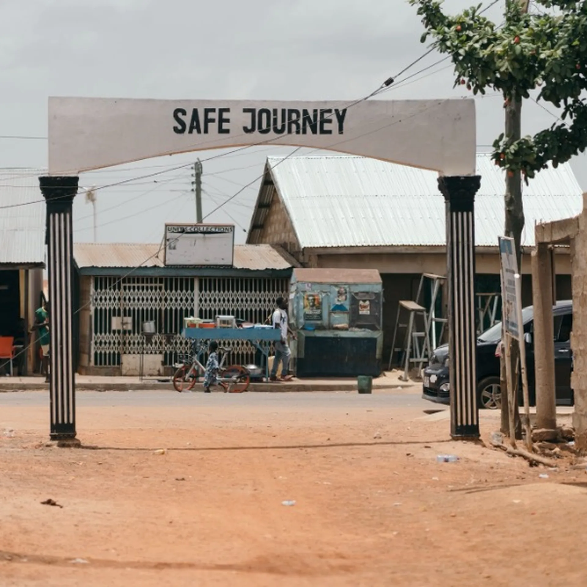 Street scene with an archway reading "Safe Journey," surrounded by small buildings, motorcycles, and people. Dirt road leads into a village.