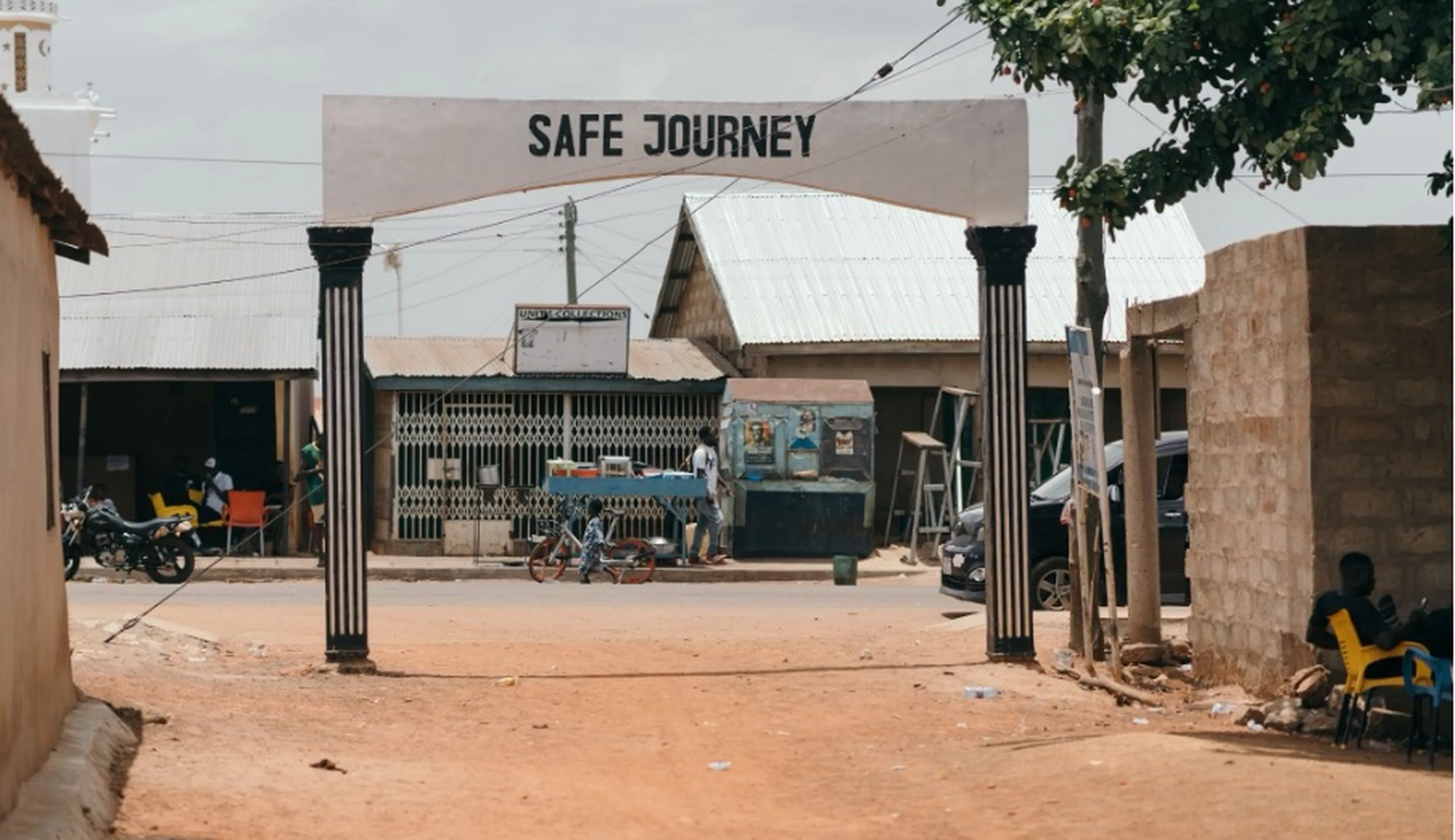 Street scene with an archway reading "Safe Journey," surrounded by small buildings, motorcycles, and people. Dirt road leads into a village.