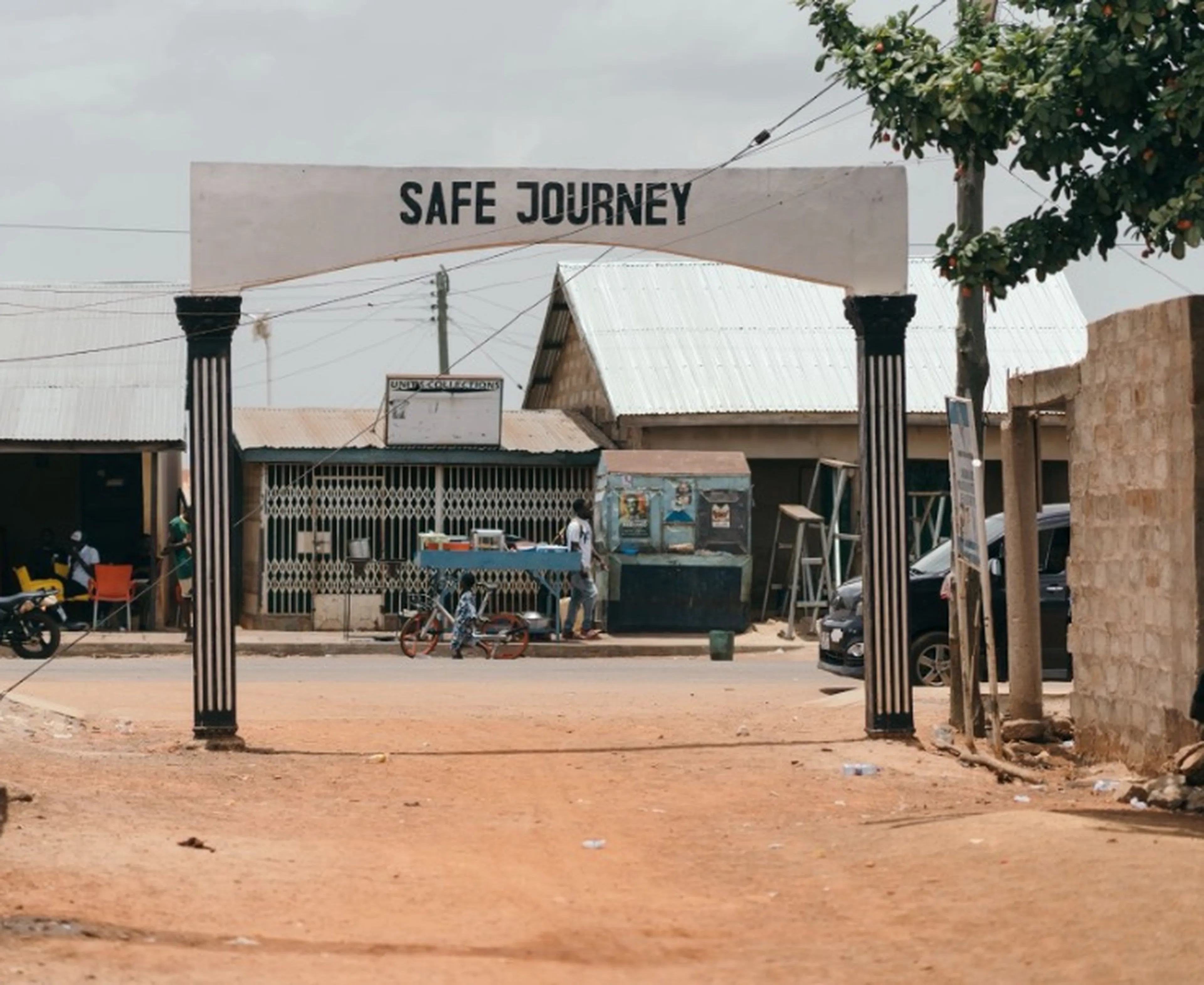 Street scene with an archway reading "Safe Journey," surrounded by small buildings, motorcycles, and people. Dirt road leads into a village.