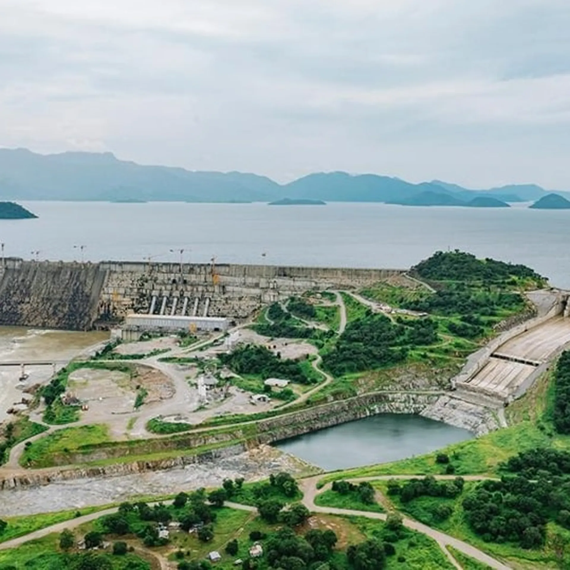 Aerial view of a large dam with multiple spillways, surrounded by lush greenery and mountains under a cloudy sky.