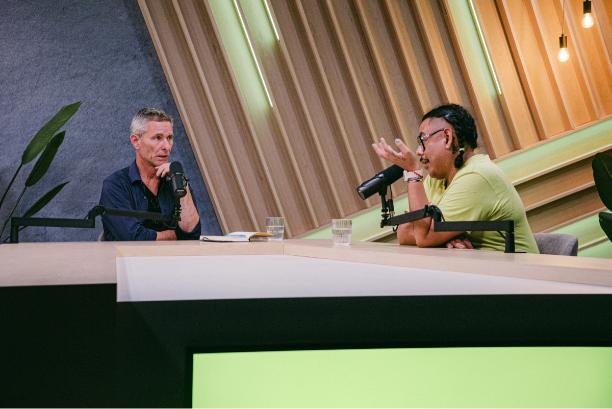 Two hosts sit at a webinar table facing each other, speaking into microphones in a modern studio with wood paneling and green accents.