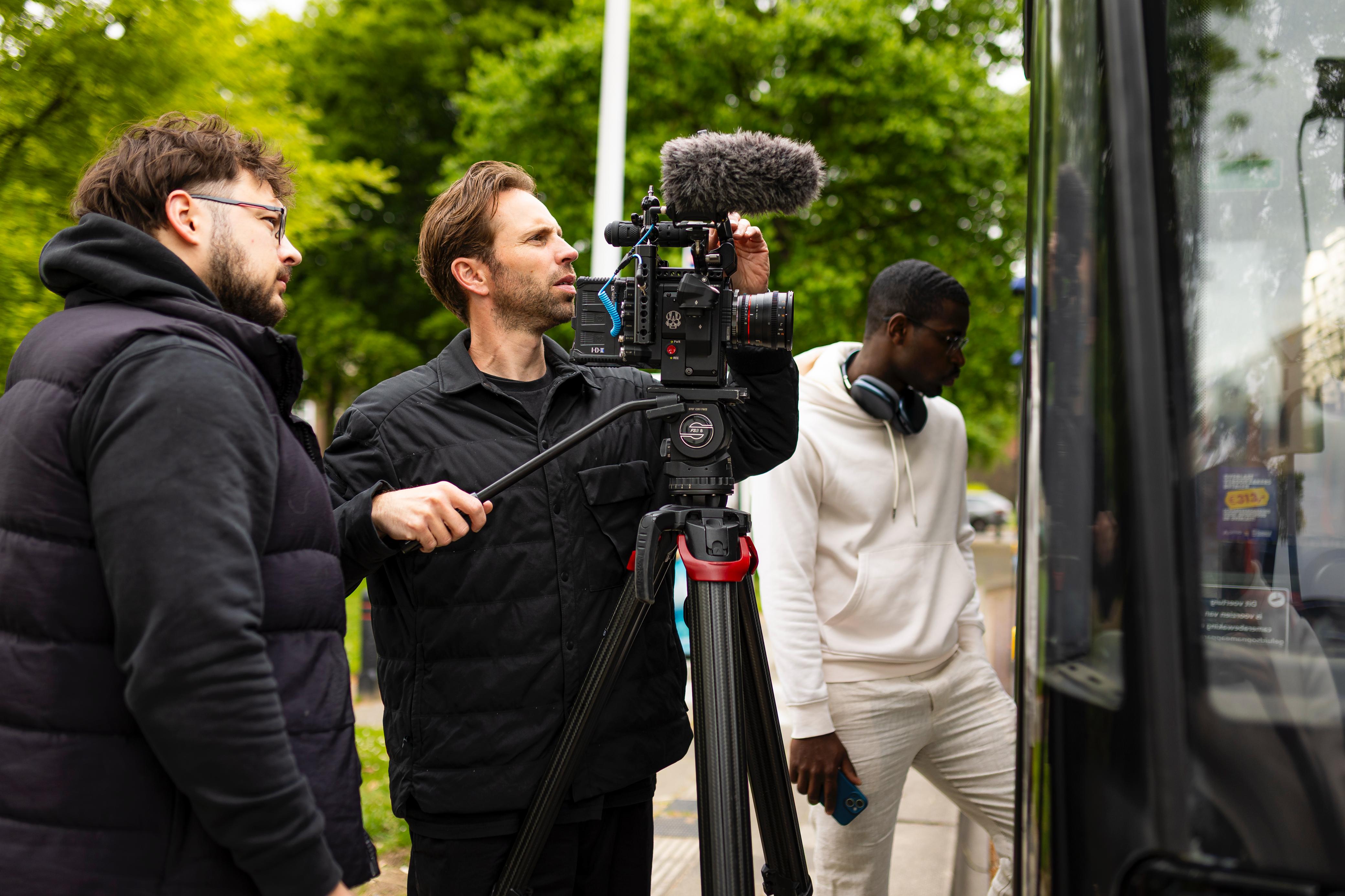Three crew members filming outdoors: one adjusts a camera on a tripod, another watches, third stands nearby with headphones and a phone.