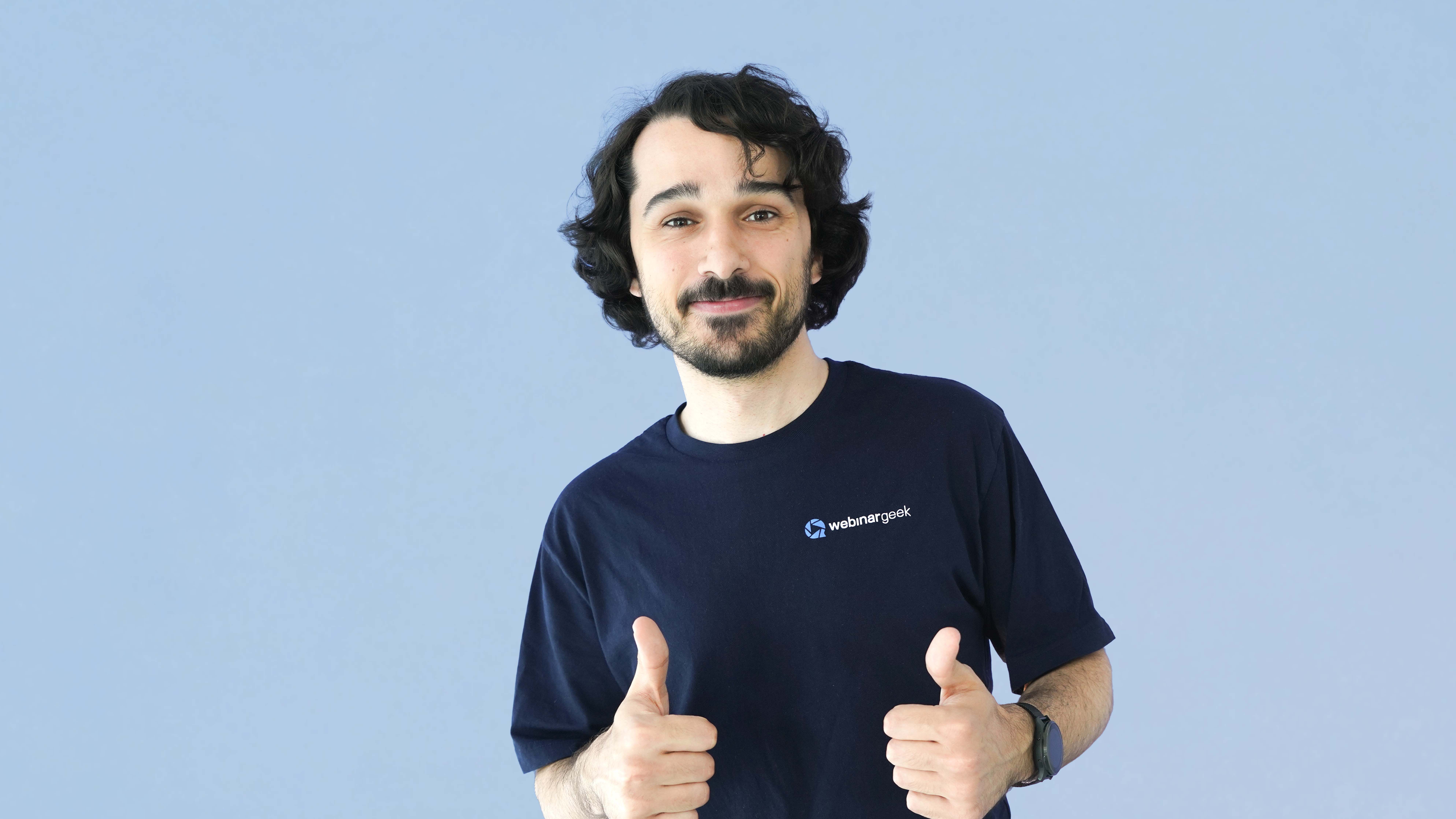 Man with curly hair and a beard, wearing a "webinargeek" T-shirt, giving two thumbs up against a plain light blue background.