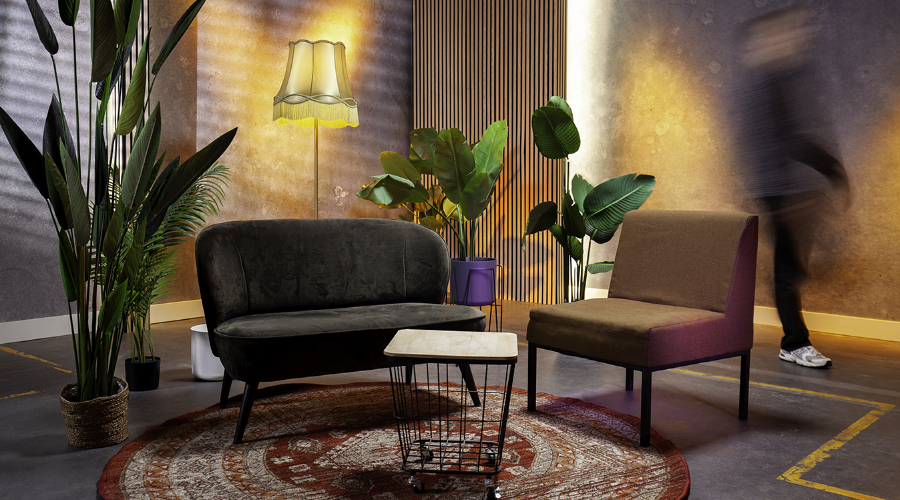 A modern living room with a black sofa, brown chair, plants, a round rug, and a vintage lamp. A blurred figure walks by on the right.