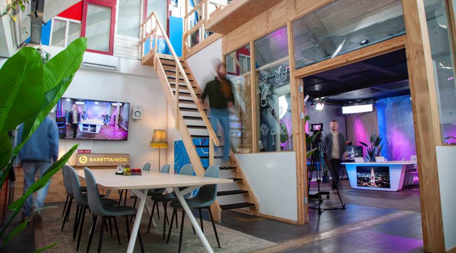 Modern office space with a central wooden staircase, dining area with chairs, large screen, and a studio area with lighting and equipment.