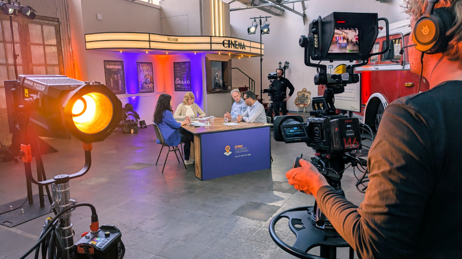 A group of people are seated at a table in a studio setting with professional lighting and cameras, with a cinema-themed backdrop.
