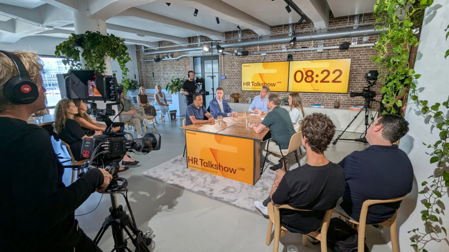 A live HR talk show with a panel of speakers seated at a table, surrounded by cameras and audience members in a modern studio setting.