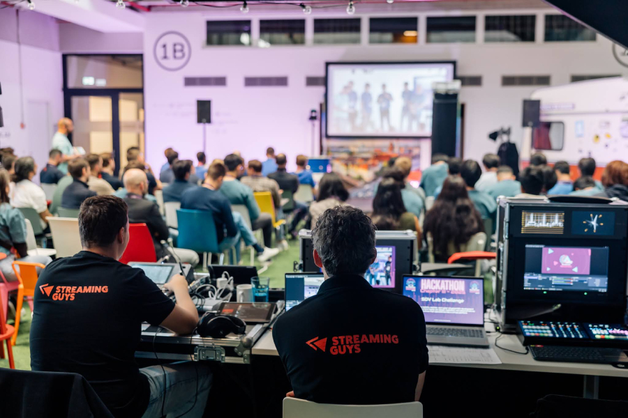 A tech event with attendees seated in a room, watching a presentation on screen. Two technicians operate equipment at the back labeled "Streaming Guys."